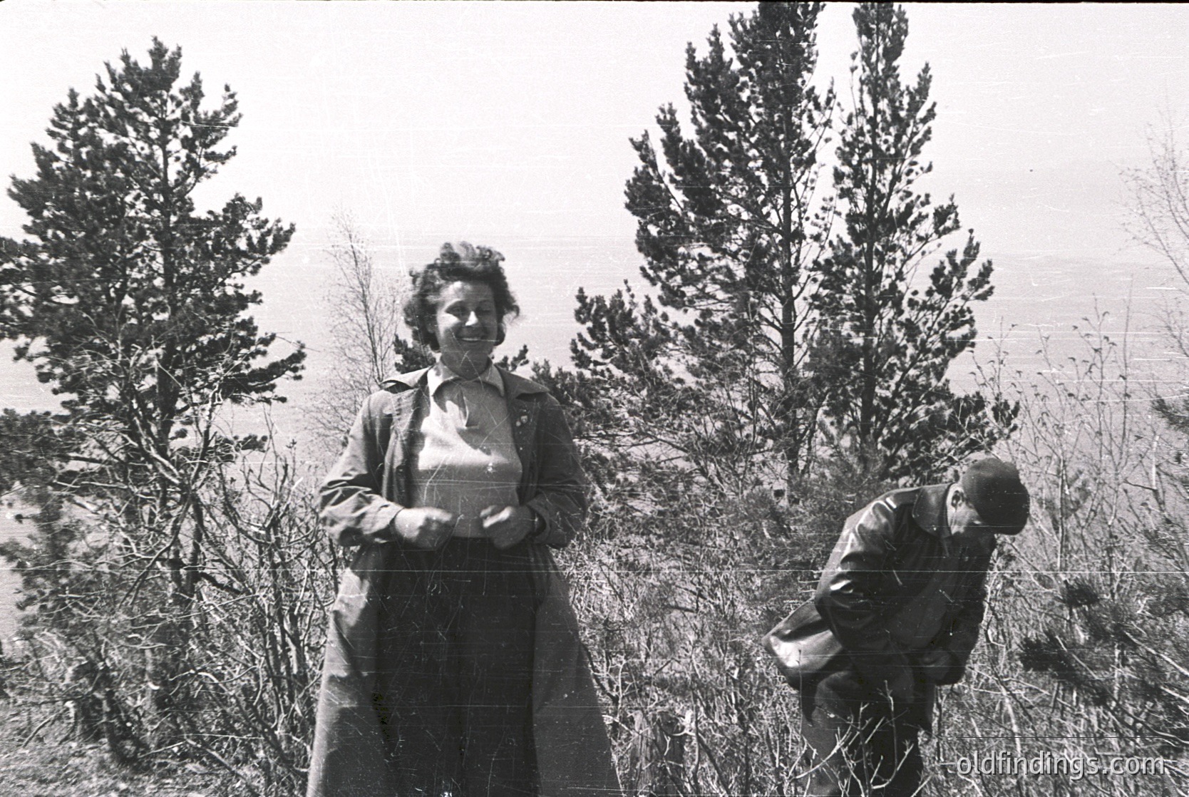 Black-and-white portrait of two individuals in a forested, hilly landscape, mid-20th century. Woman in long coat and headscarf stands upright, holding a small object; man in cap and coat leans forward, possibly picking berries or flowers. Dense pine trees frame the scene, suggesting rural or mountainous terrain.