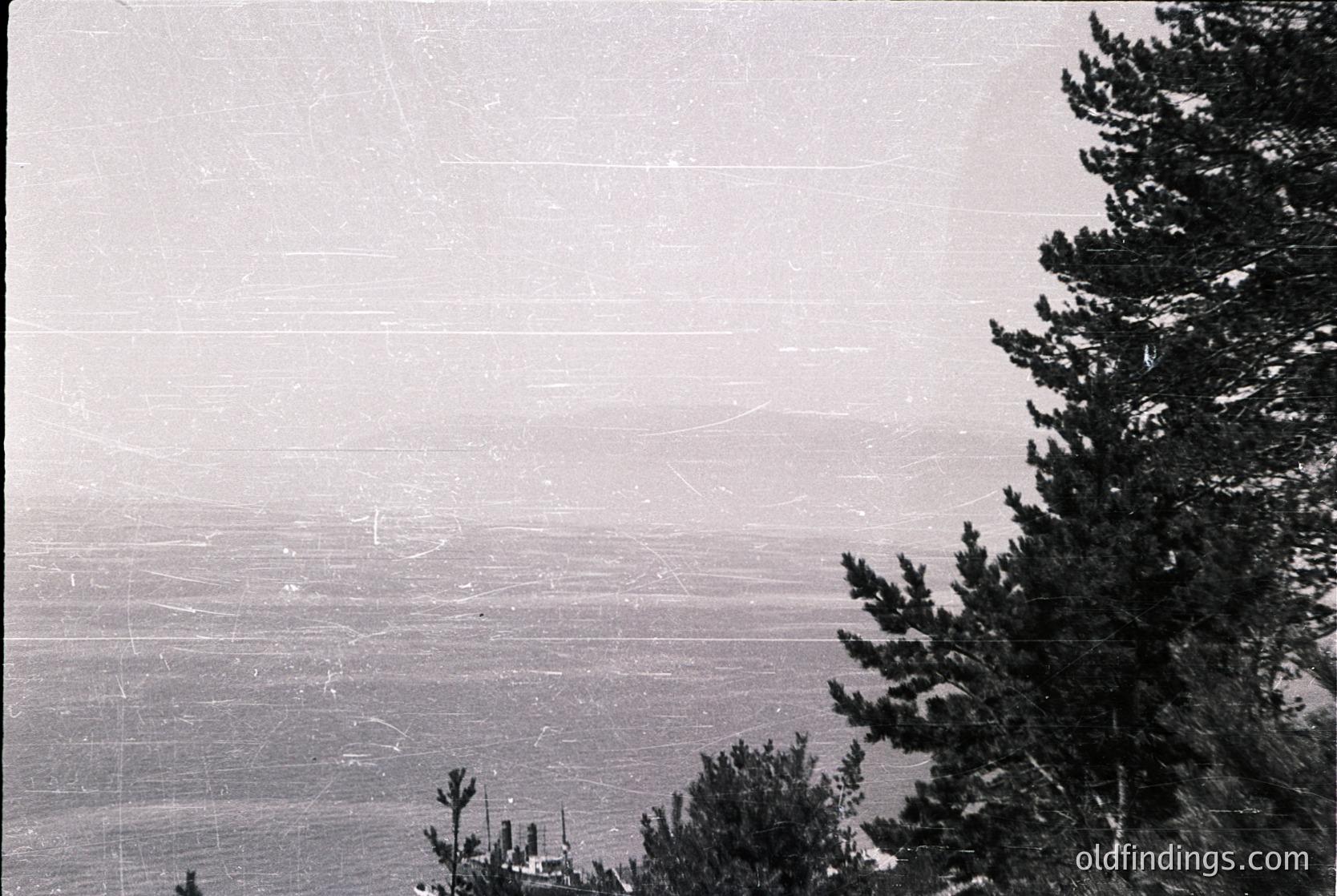 Vintage aerial view of forested mountain ridge with misty valley below. Coniferous trees frame the right edge; faint road or trail winds through lower terrain. Likely mid-20th century due to grainy, high-contrast film style.