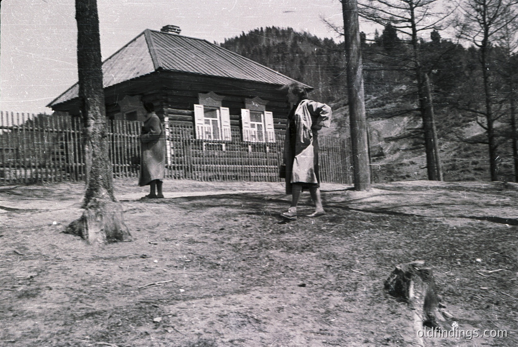 Two individuals in traditional alpine attire stand near a rustic wooden cabin with a peaked roof, framed by a simple metal fence. One holds a bundle, while a dog rests in the foreground. Forest and rocky terrain in the background suggest a mountainous region.