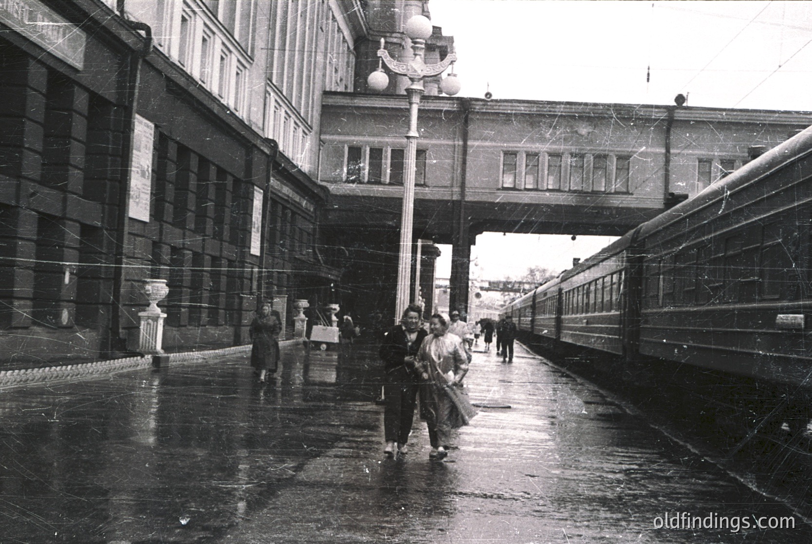 Mid-20th century train station platform with wet pavement, featuring Soviet-era architecture. A group of people in 1950s-60s attire—men in suits, women in long skirts—walk toward a parked passenger train. Overhead bridge and decorative lampposts frame the scene.
