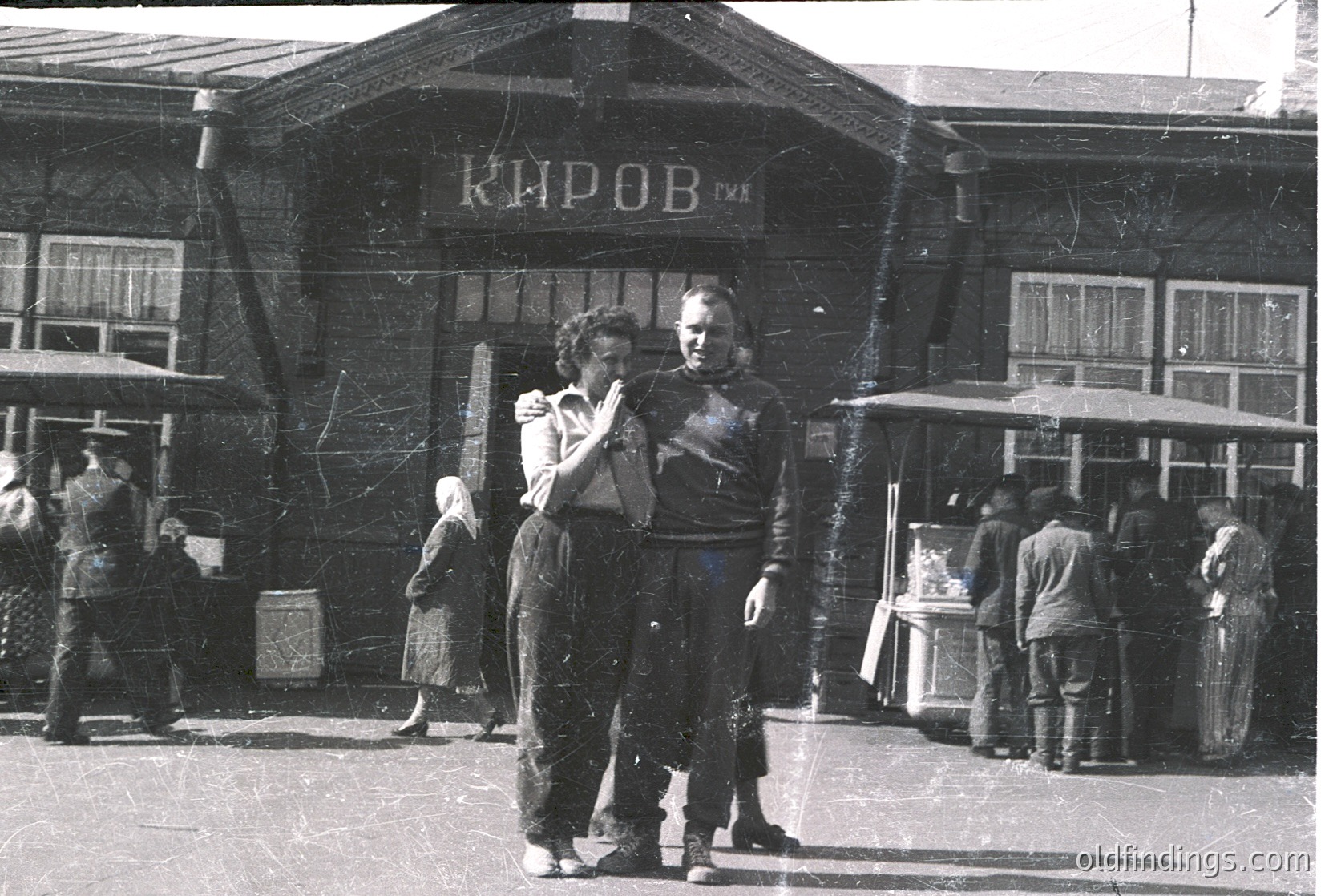 Couple dancing near a Soviet-era "Киров" (Kirov) building, likely a train station or cultural center, in mid-20th century. Wooden architecture with Cyrillic signage and vintage clothing (1950s–1960s). Urban street scene with pedestrians and a kiosk.