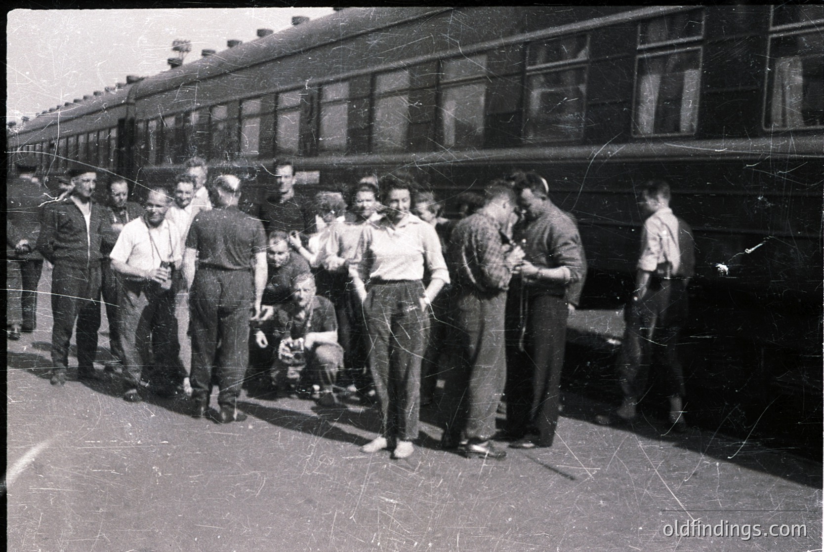 Vintage black-and-white photo of a group of men posing near a mid-20th-century passenger train, likely 1950s–1960s. Uniform workwear (overalls, caps) suggests industrial or agricultural background. Train features classic riveted metal construction and large windows. Station platform appears utilitarian, with visible tracks and overhead wires.