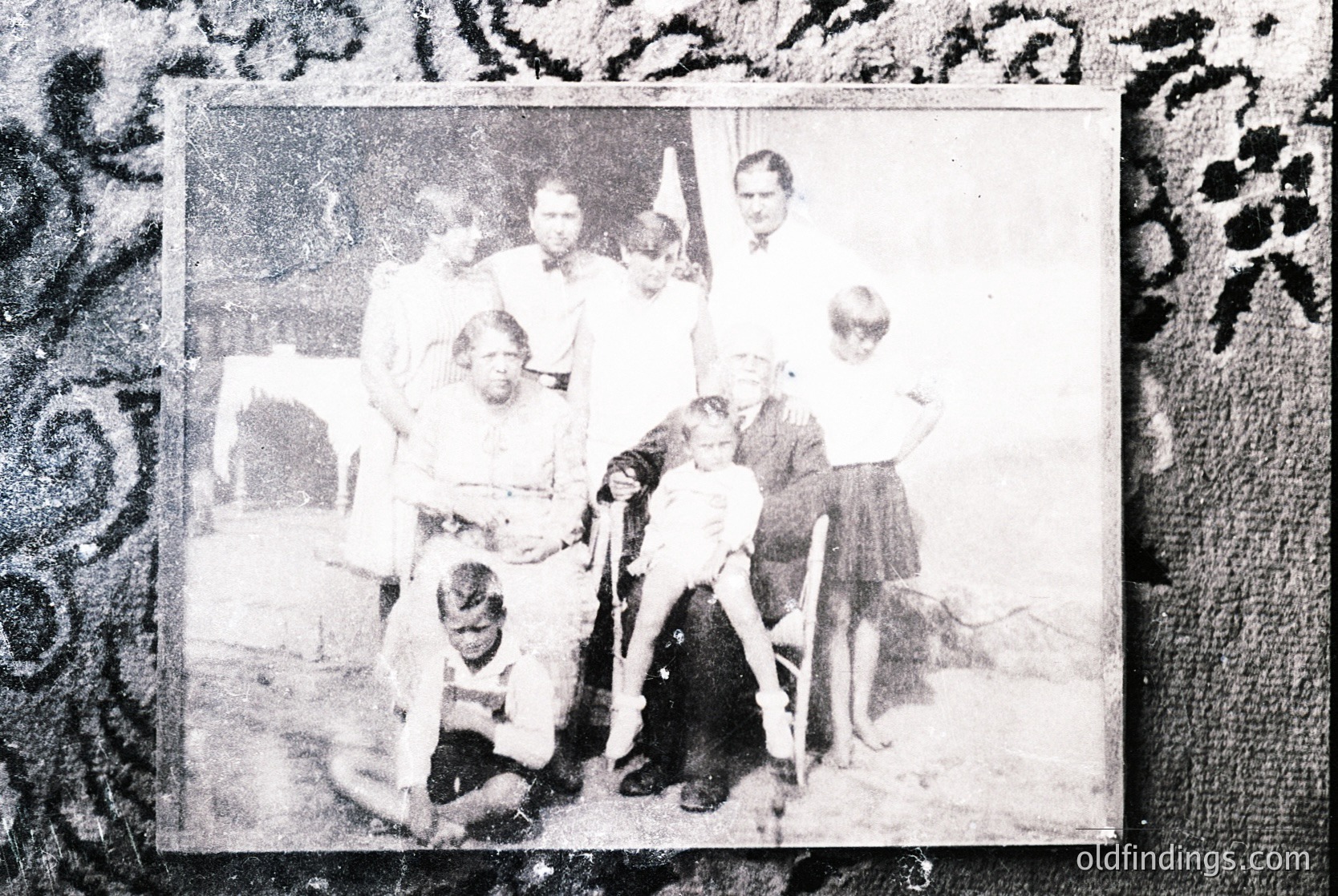 Vintage black-and-white family portrait from the early-to-mid 20th century, likely 1940s–1950s. Nine individuals pose indoors, framed by ornate decorative border. Adults wear formal attire (suits, dresses), children in casual clothing. Indoor setting suggests a living room with visible furniture.