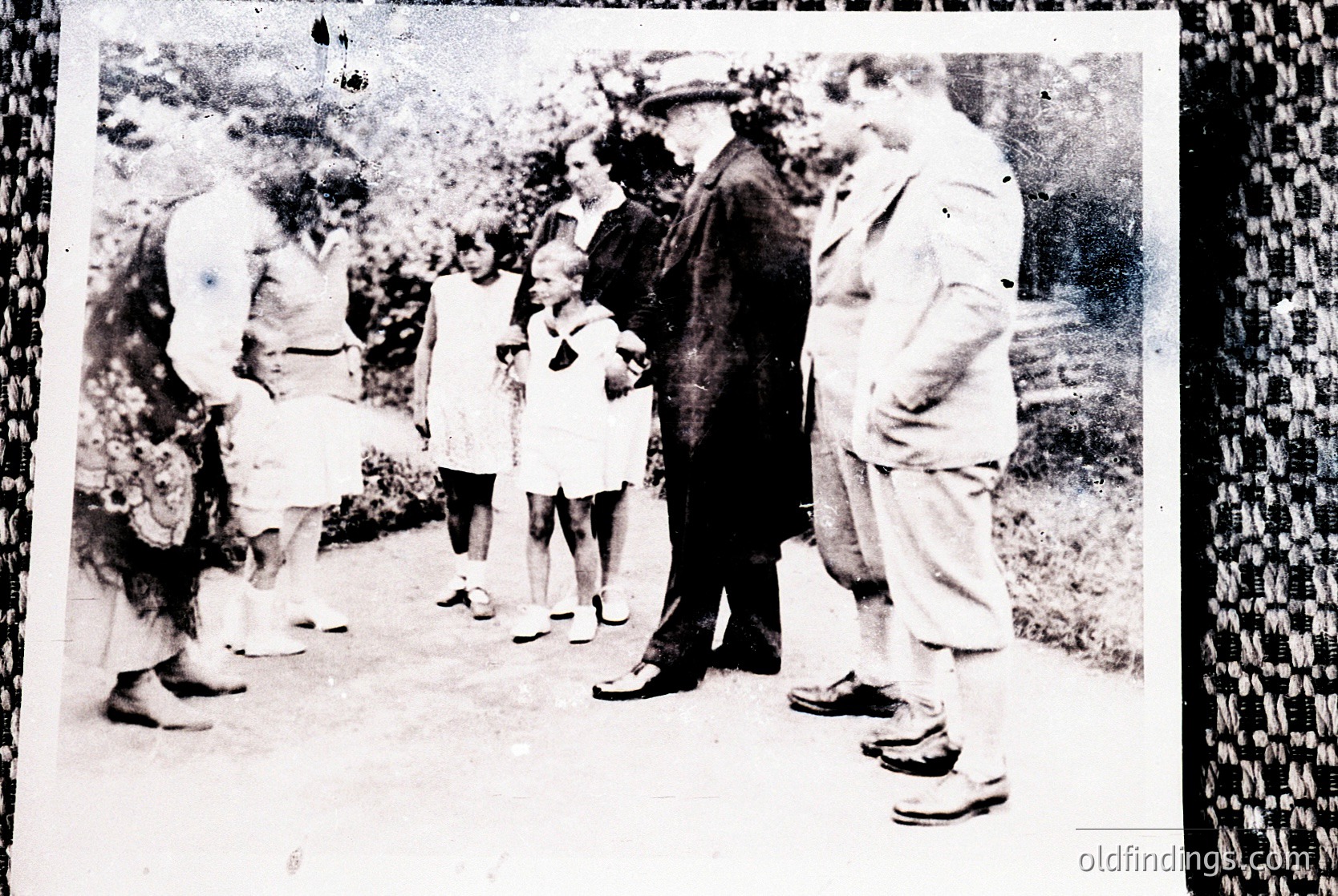 Vintage black-and-white photo of a rural gathering: adults in loose, early 20th-century clothing (long skirts, suspenders, wide-brim hats) and children in simple dresses. Group stands on a dirt path beside lush greenery, suggesting a communal or family event. Likely Eastern European or rural American setting, 1910s–1930s.