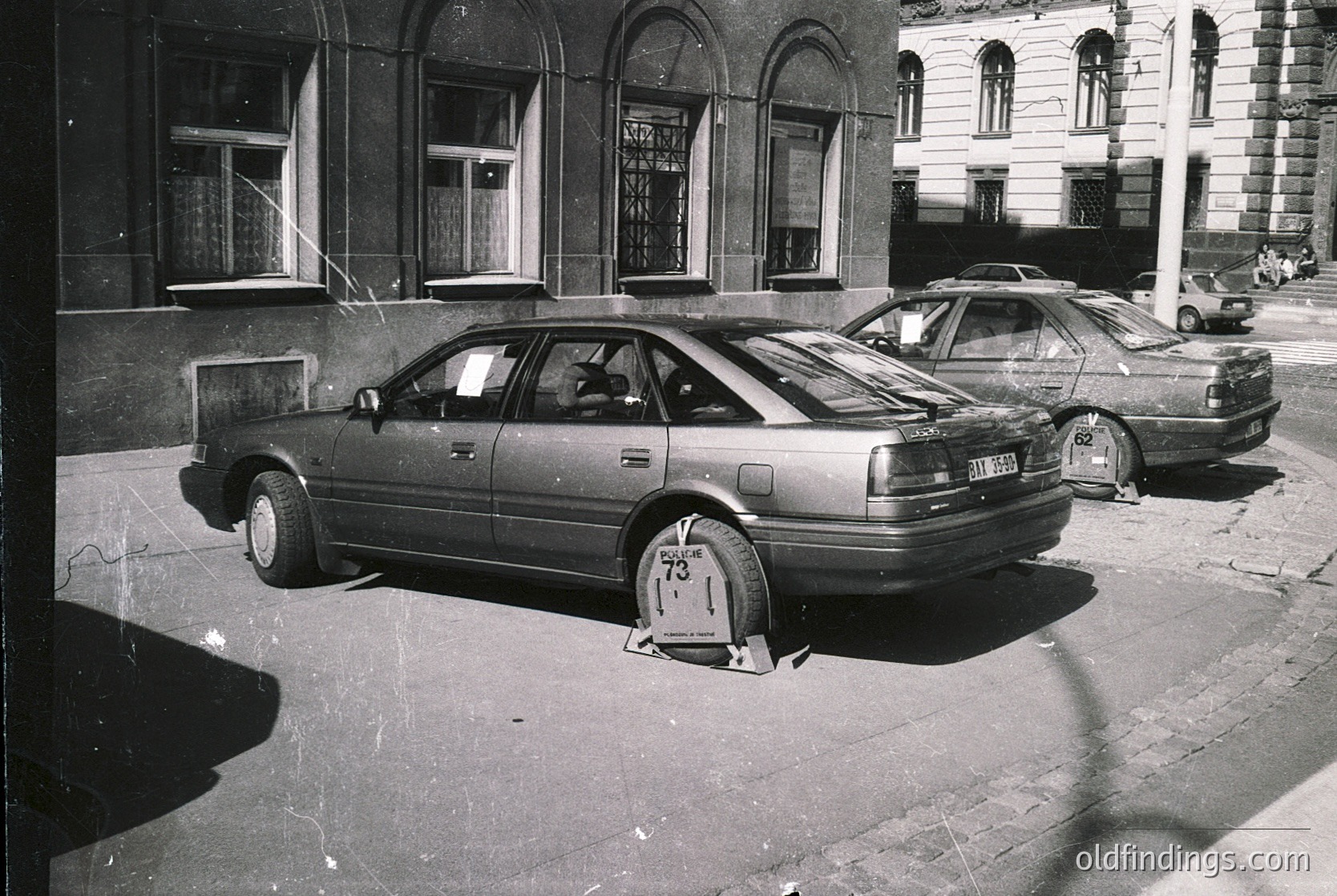 1970s-era sedan parked on cobblestone street with Soviet-style parking block. Neo-classical building with arched windows and shuttered ground floor in urban setting. Vintage European cityscape with minimalist signage.
