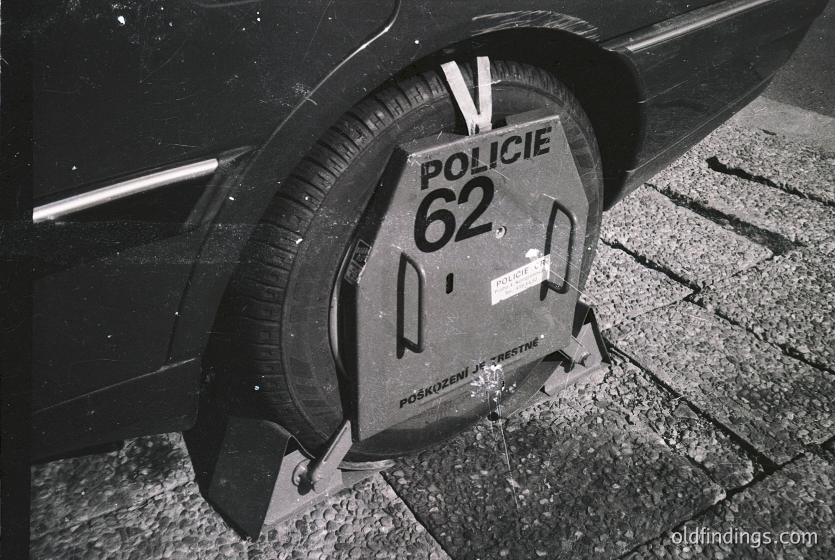 Black-and-white close-up of a Polish police wheel clamp labeled "Policja 62" on a vehicle tire, indicating a parking violation. The clamp bears Cyrillic text ("POZKOZENIE JESTE") and a warning sticker. Likely Eastern Europe, mid-to-late 20th century.