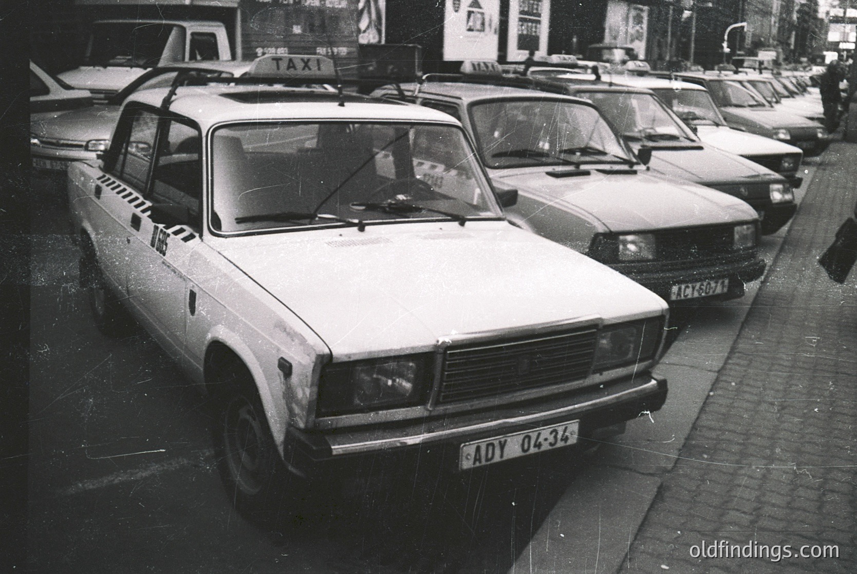 Vintage taxi fleet in urban setting, featuring a prominent Lada taxi (ADY 04-34) with "TAXI" signage and black-and-white checkered roof. Other Soviet-era sedans parked in a row, likely Eastern Bloc city streets. Mid-20th century urban transport, indicative of 1970s–1980s Eastern Europe.