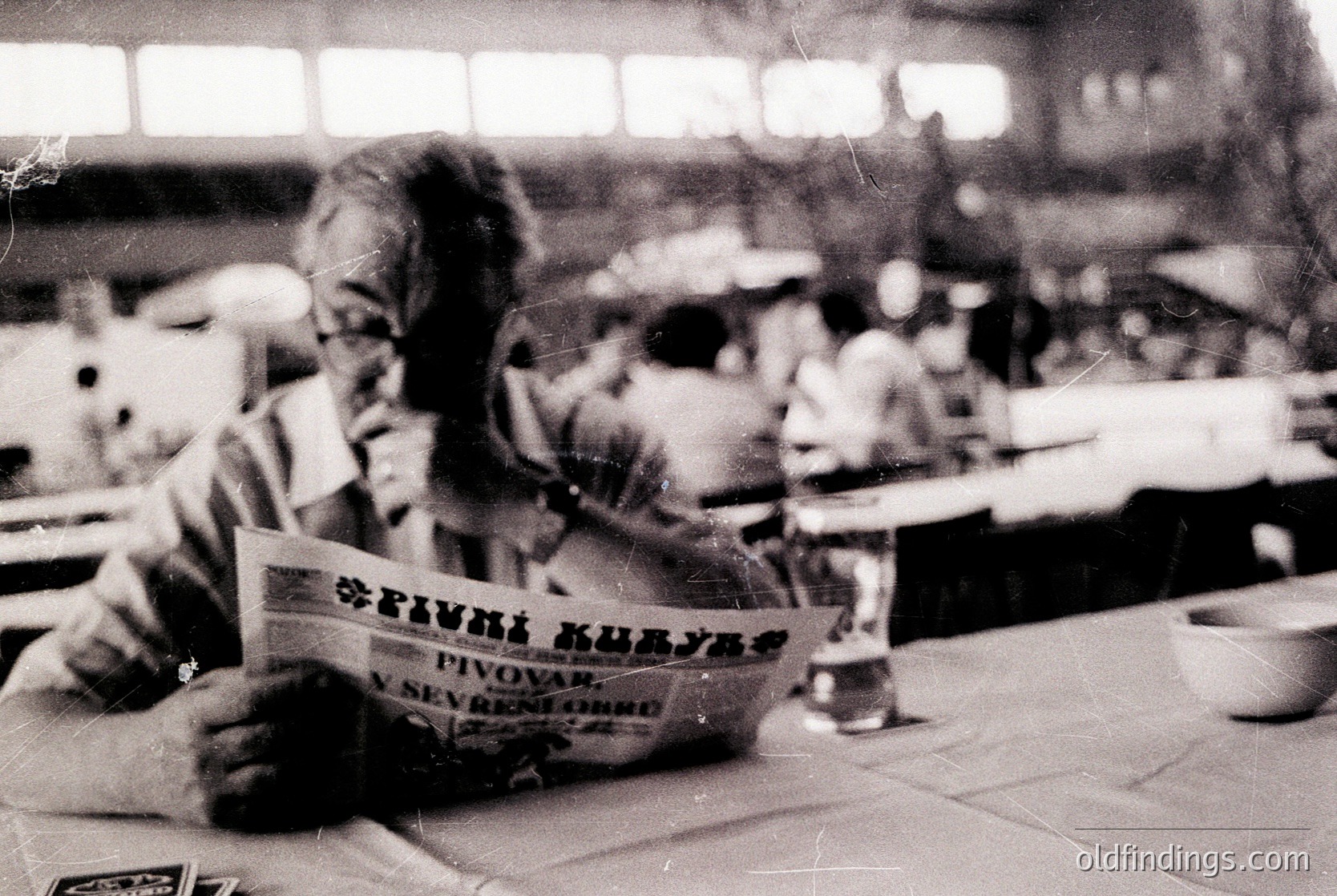 Vintage black-and-white photo of a man reading a Bulgarian newspaper ("Пиво Куявец" by Pivovар "Sevtopore") in a café or restaurant, likely from the 1960s–70s. Striped shirt and glasses suggest mid-century fashion; blurred background shows tables, chairs, and patrons. Ideal for historical research or nostalgic design references.
