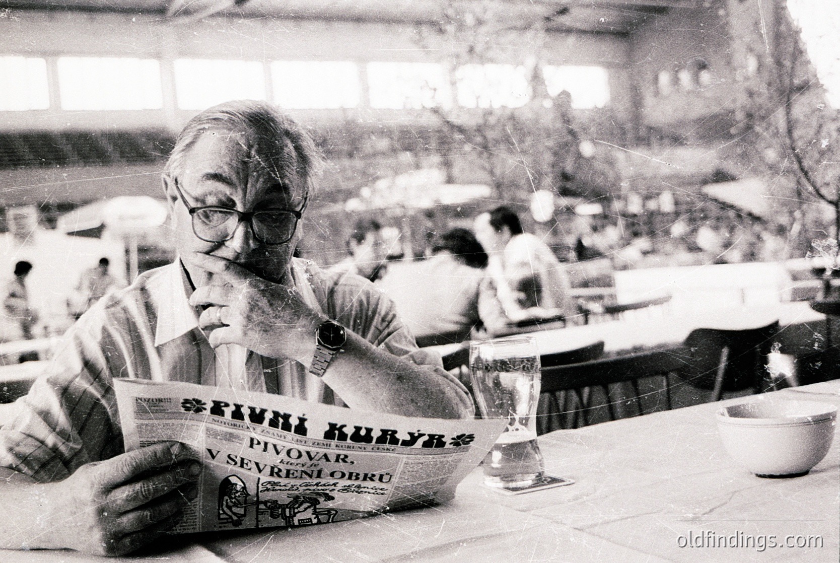 Mid-20th century café scene: Man in striped shirt reads *Pivni Kuliyar* beer menu (Bulgarian: "Beer Tavern") at a seaside or resort-style dining hall. Glasses, plates, and blurred patrons suggest a lively, communal atmosphere. Likely – Bulgaria ( or Black Sea coast).