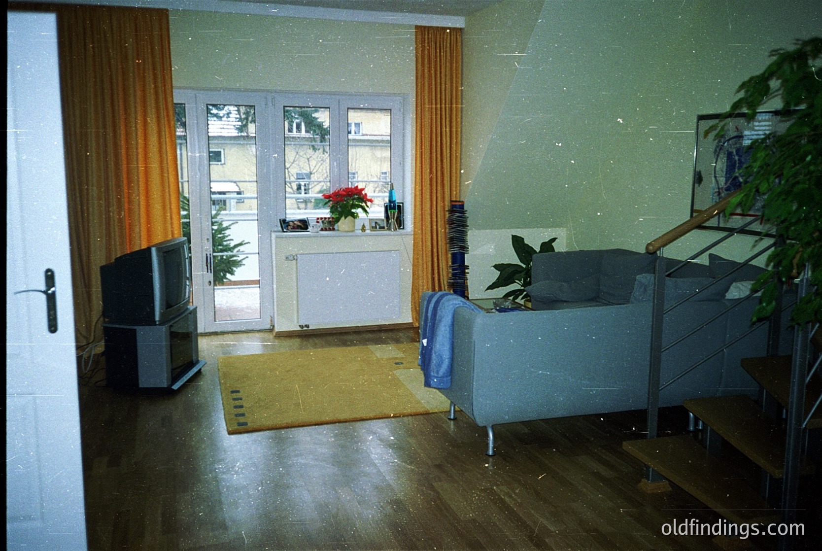 Vintage living room featuring mid-century design elements: orange curtains framing a sliding glass door, a small CRT TV on a wooden stand, and a sofa with metal legs. Decor includes a potted poinsettia by the window and a potted plant on a side table. Light-colored linoleum flooring and a yellow doormat at the entrance. Likely Eastern Bloc-era interior, 1970s–1980s.