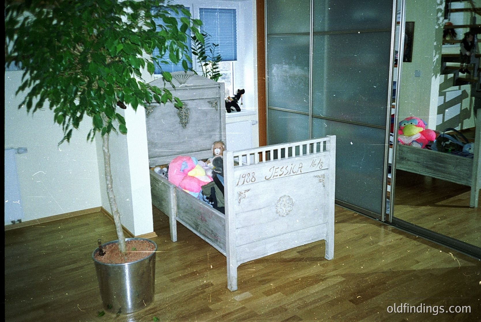 Vintage wooden crib (1928) with "Jessie" engraved, positioned under a potted plant in a dimly lit room. Wooden floor and metal bucket on left. Blanket and stuffed toy on crib. Reflective glass doors and shelving in background. Likely mid-20th century domestic setting.