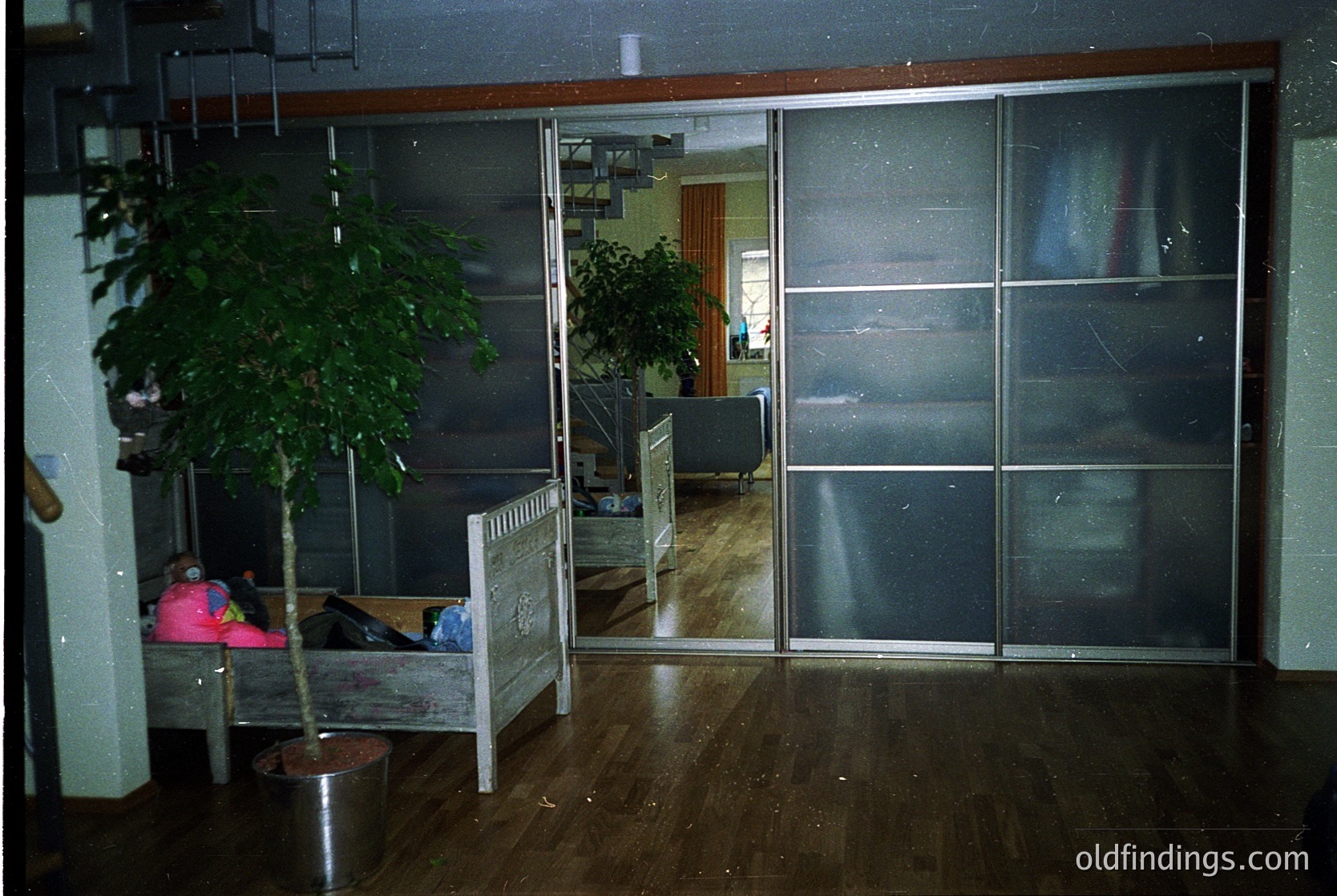 Mid-century modern interior with sliding glass partitions, reflecting a minimalist office or waiting area. Wooden floor, potted plants, and a child’s chair with a pink blanket suggest a functional, utilitarian space. Reflections reveal a sofa and desk in the adjacent room. Likely – design.