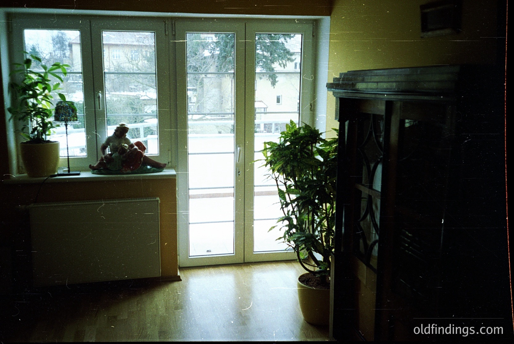 Mid-century interior with sliding glass doors revealing a snowy courtyard. A child sits on a built-in bench beside a potted plant, while a wooden cabinet and large potted plant frame the doorway. Warm lighting contrasts with the cold exterior. Likely Eastern European residential architecture, 1970s-1980s.