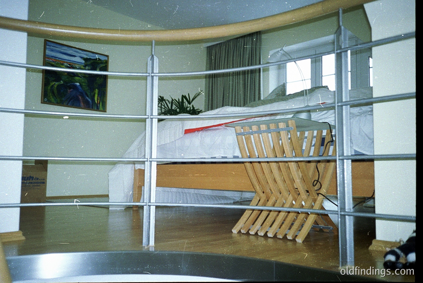 Mid-century modern hotel room with curved balcony railing. Wooden lounge chair and ottoman positioned outside, overlooking a minimalist interior with a framed abstract painting and potted plant. Boxed items suggest recent unpacking or relocation. Likely or design, indicative of functionalist aesthetics.