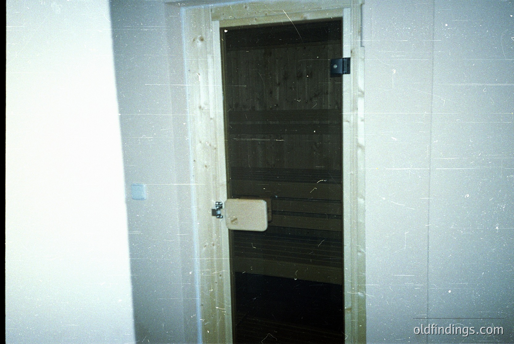 Vintage institutional door with wooden plank interior and metal latch, set in white-tiled corridor. Likely mid-20th century institutional or medical facility.