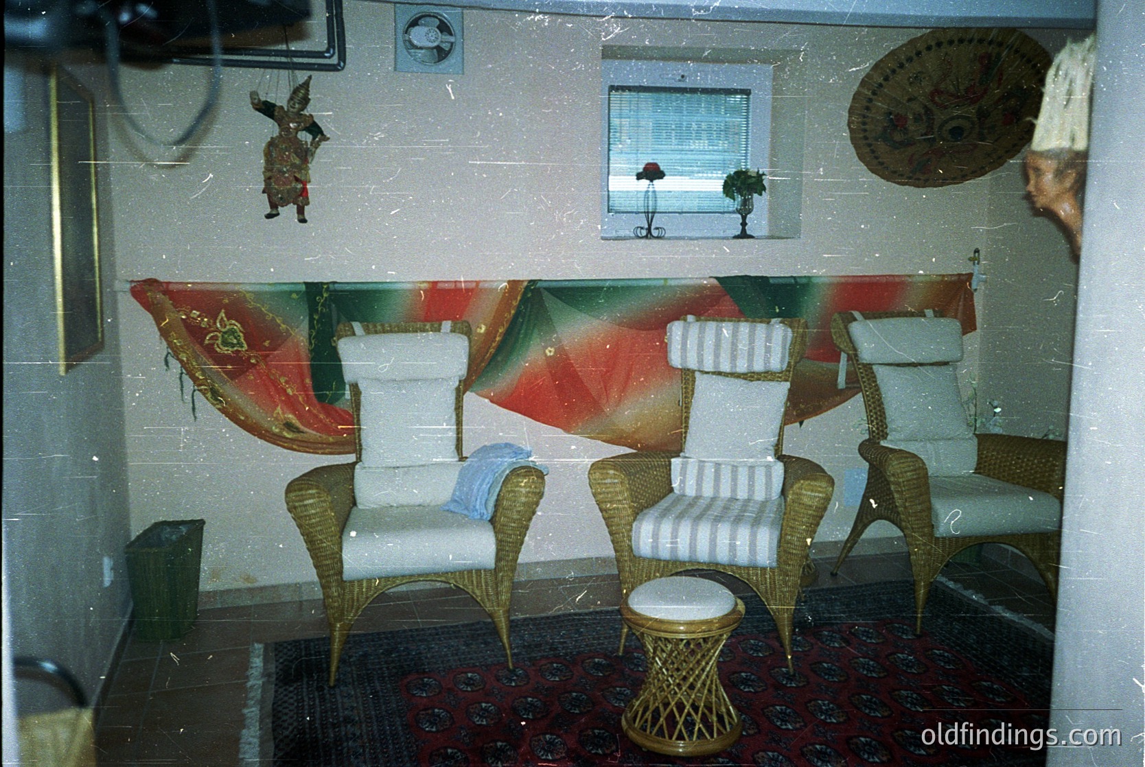 Vintage indoor seating area with mid-century modern wicker chairs, striped cushions, and a woven stool. Bold geometric wallpaper features red, green, and white patterns. Decorative wall art includes a hanging marionette and framed tropical scene. Reflective surface suggests a glass barrier or door. Likely 1960s–1970s interior design.