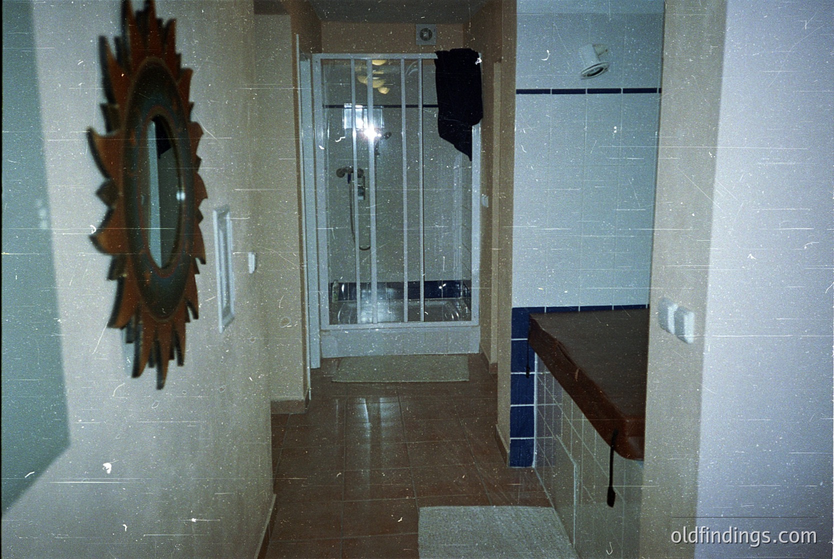 Mid-century bathroom with tiled walls in neutral tones, featuring a sunburst-style mirror on left. Glass-block door with metal frame leads to an outdoor balcony. Built-in countertop with blue-tiled edge. Lighting suggests vintage design, likely 1960s–1980s.