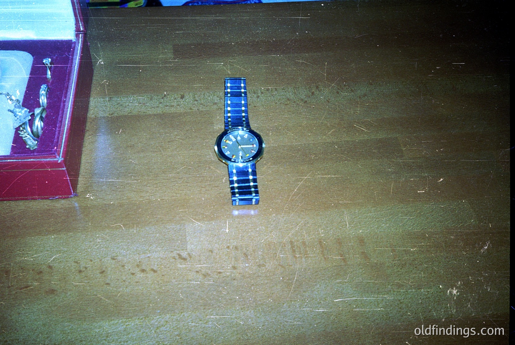 Vintage wristwatch with blue and white checkered strap, placed on worn wooden surface. Circular dial with minimalist design, likely from the 1970s–1980s. Partial view of a red gift box in background suggests retail or display setting.