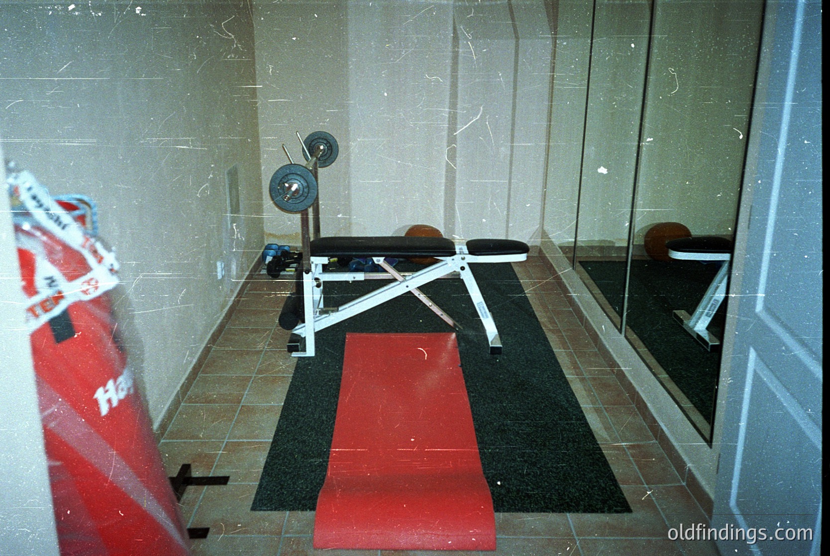 Vintage gym bench press station in a tiled, utilitarian space. White adjustable bench with weight stack, red rubber mat, and mirrored wall reflecting equipment. Likely 1980s–1990s Soviet/Eastern Bloc gym design. Functional, minimalist fitness setup.