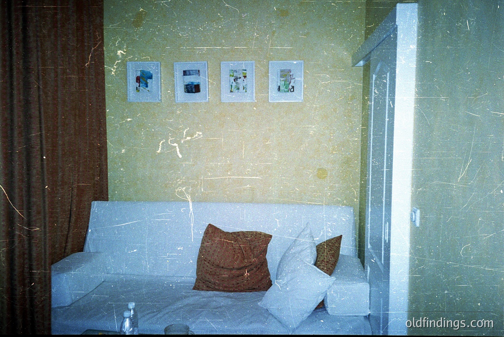 Vintage hotel room with mid-century design—marble-walled corridor leading to a sofa bed in muted tones, framed photos depicting coastal scenes. Glass door reveals a glimpse of a hallway.