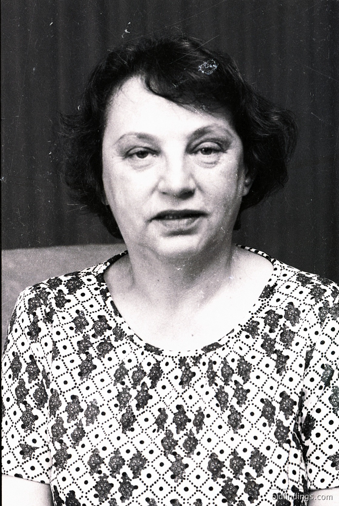 Portrait of an individual in a patterned blouse with floral and geometric motifs, likely mid-20th century. The dark, plain background suggests studio or formal portraiture. Hair styled in a short, rounded bob with bangs.