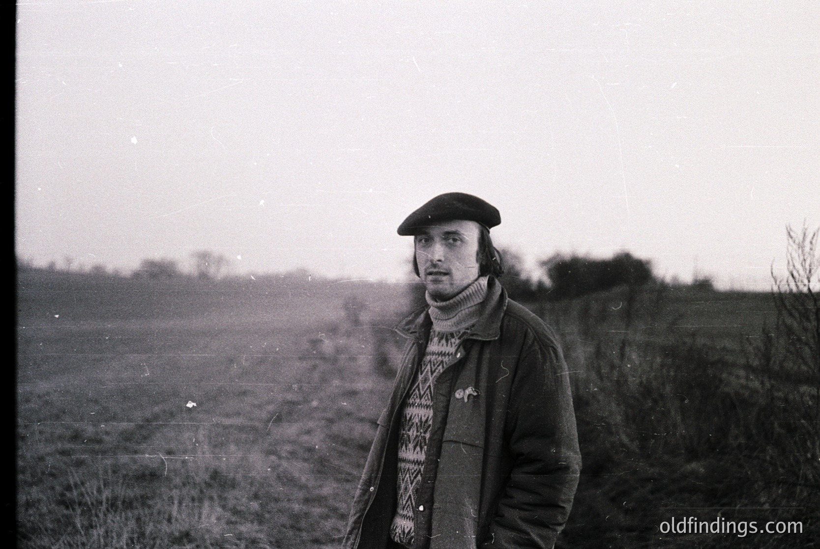 Black-and-white portrait of a man in a rural landscape, wearing a beret, plaid scarf, and a long coat. His gaze is directed toward the camera, suggesting a candid yet composed moment. The blurred background reveals open fields and sparse vegetation, indicative of a countryside setting. The grainy texture and lighting suggest a mid-20th century photograph, likely from the 1950s–1970s. [Vintage portrait of a man in countryside ]