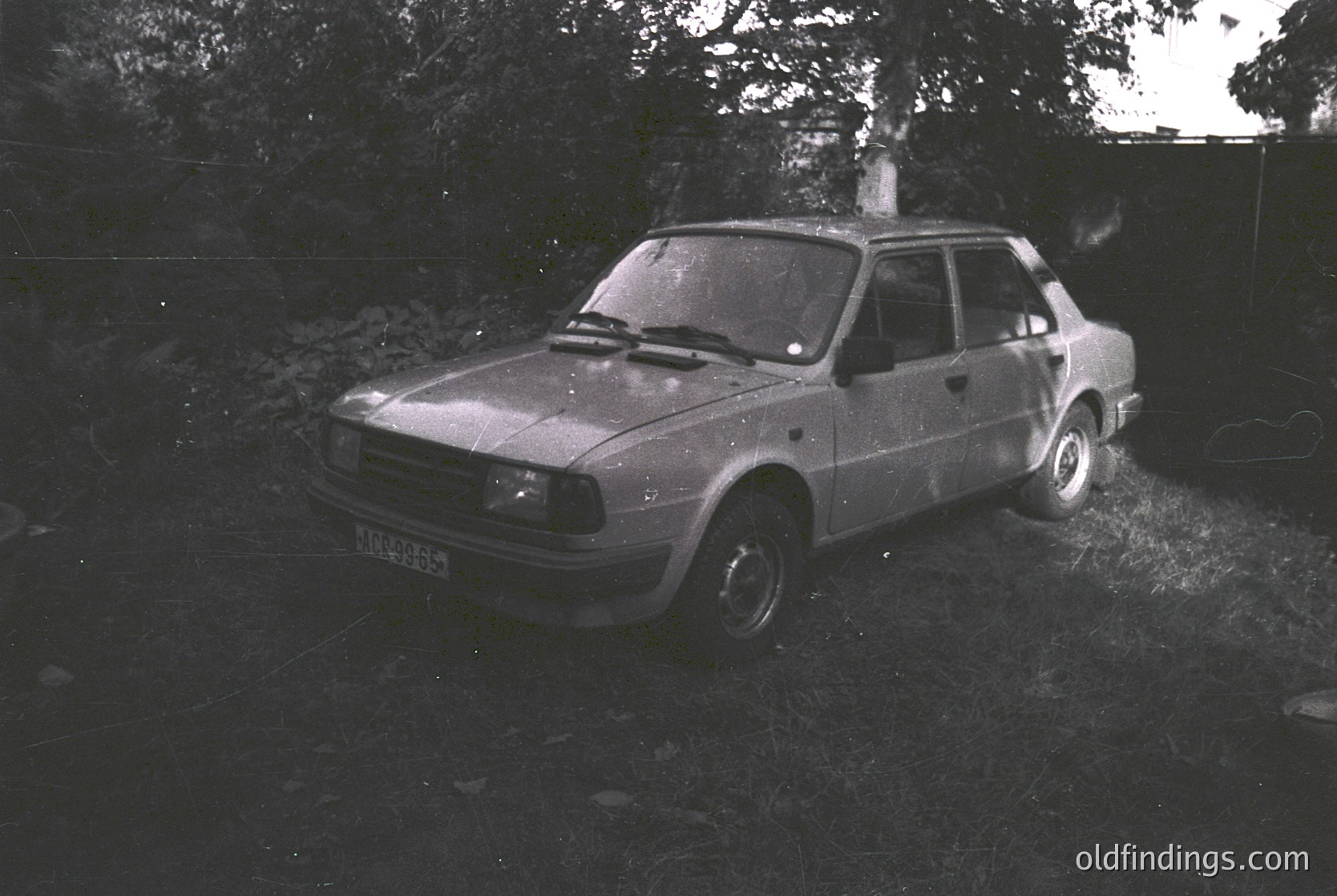 Vintage hatchback sedan parked on uneven grass beside a stone wall, likely a Škoda from the 1970s–1980s. Distinctive boxy design with rounded corners and minimalist styling. Blurred background suggests motion or low-light exposure. Ideal for automotive history, nostalgia, or stock photography.