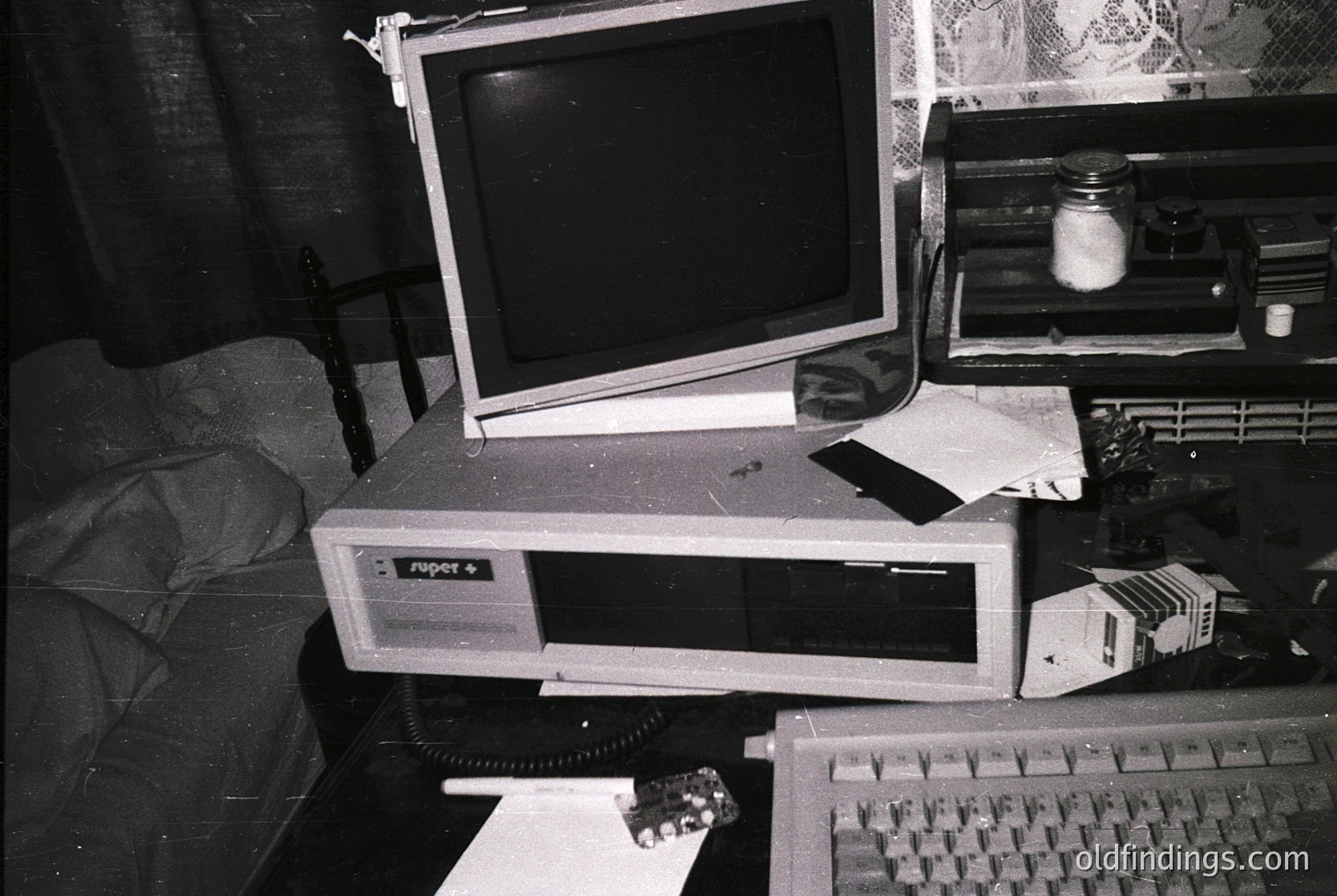 Retro desktop setup featuring a **Commodore Amiga 500** (front) with CRT monitor, keyboard, and mouse. Surrounding clutter includes paper stacks, a small glass container, and a patterned curtain. Evokes late **1980s–early 1990s** home computing nostalgia.