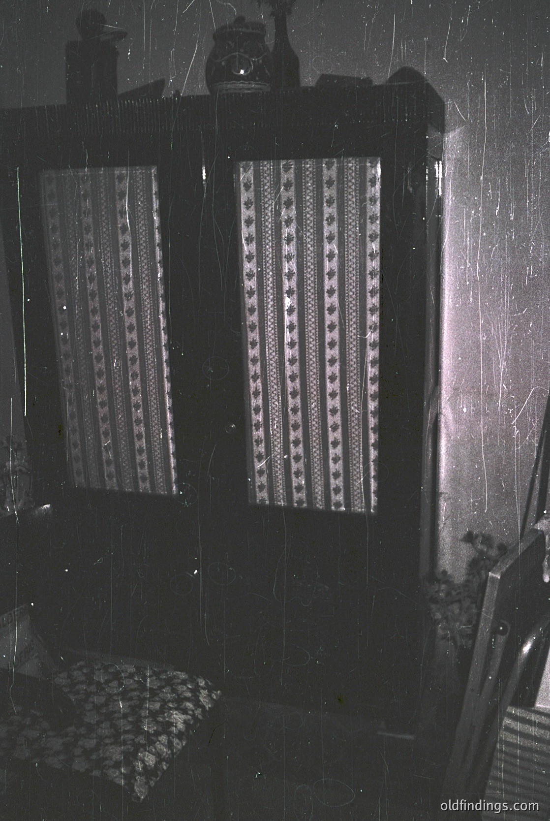 Vintage interior featuring patterned curtains with geometric designs, likely from the 1960s–1970s. Dark wood-framed windows with visible rain streaks, paired with a patterned cushion and floral wallpaper. Industrial-era lighting fixtures and a partial view of a wicker chair. Ideal for historical home decor or mid-century design research.