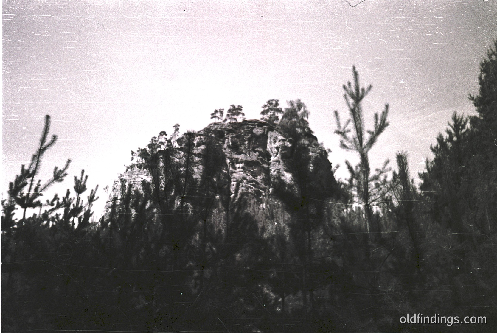 Monochrome landscape featuring a jagged rock formation rising from dense forest. The rugged peak contrasts with sparse, leafless trees, suggesting late autumn or early spring. Vintage grain and lighting suggest mid-20th century photography.