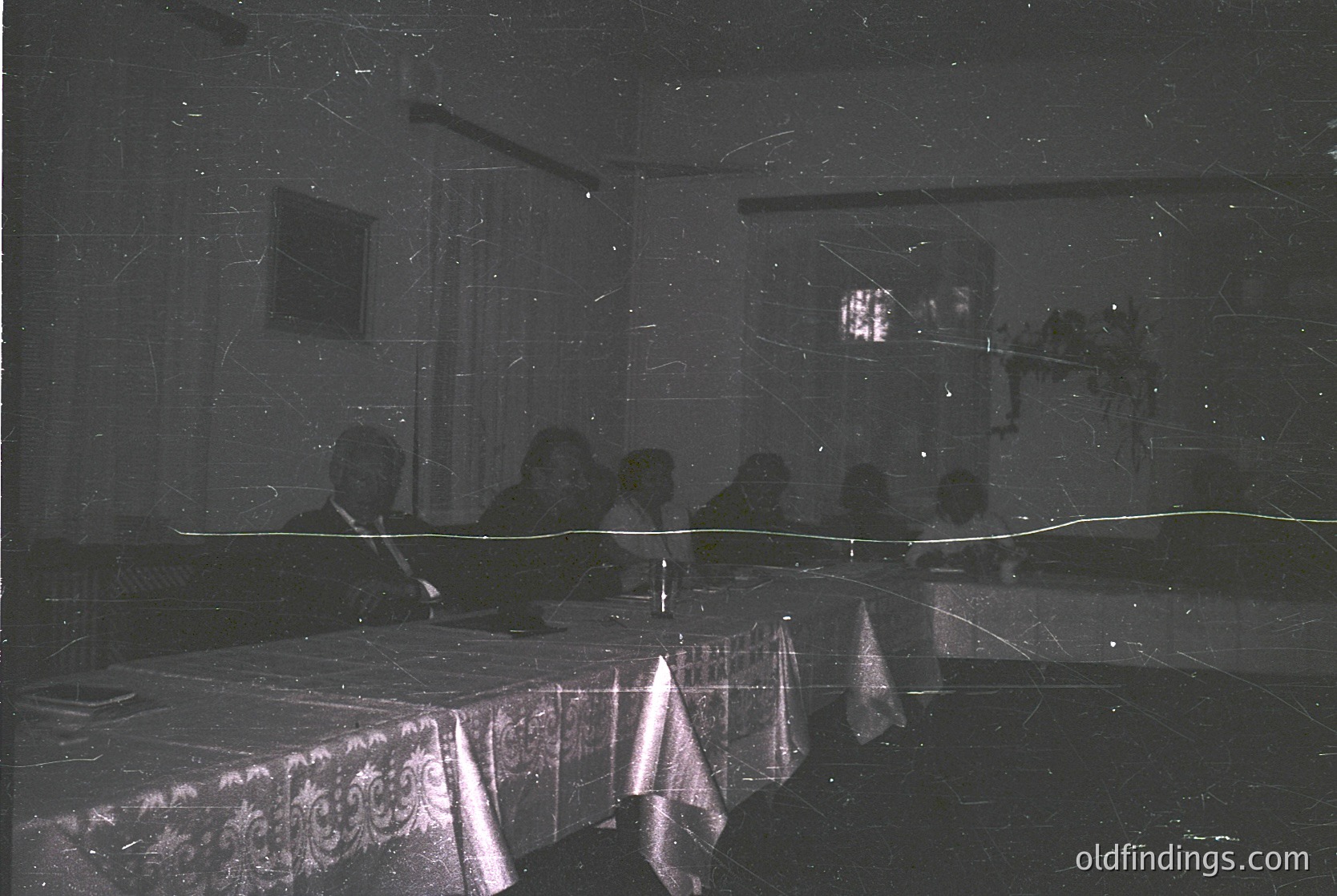 Vintage black-and-white photo of a dimly lit indoor gathering, likely a 1960s–1970s event. Round table draped in ornate tablecloth, surrounded by blurred figures seated in chairs. Reflections and light streaks suggest motion or flash photography. Formal setting with patterned wallpaper and framed artwork. Evokes mid-century social or ceremonial atmosphere.