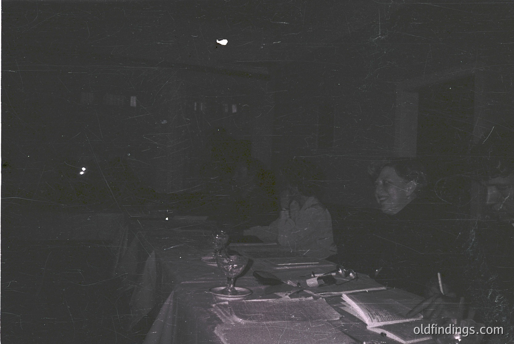 Vintage black-and-white shot of a dimly lit indoor dining scene, likely mid-20th century. Table set with white tablecloth, crystal glasses, and cutlery. Two seated figures in shadowed profiles, suggesting formal or semi-formal gathering. Dust specks and film grain enhance nostalgic vintage aesthetic.