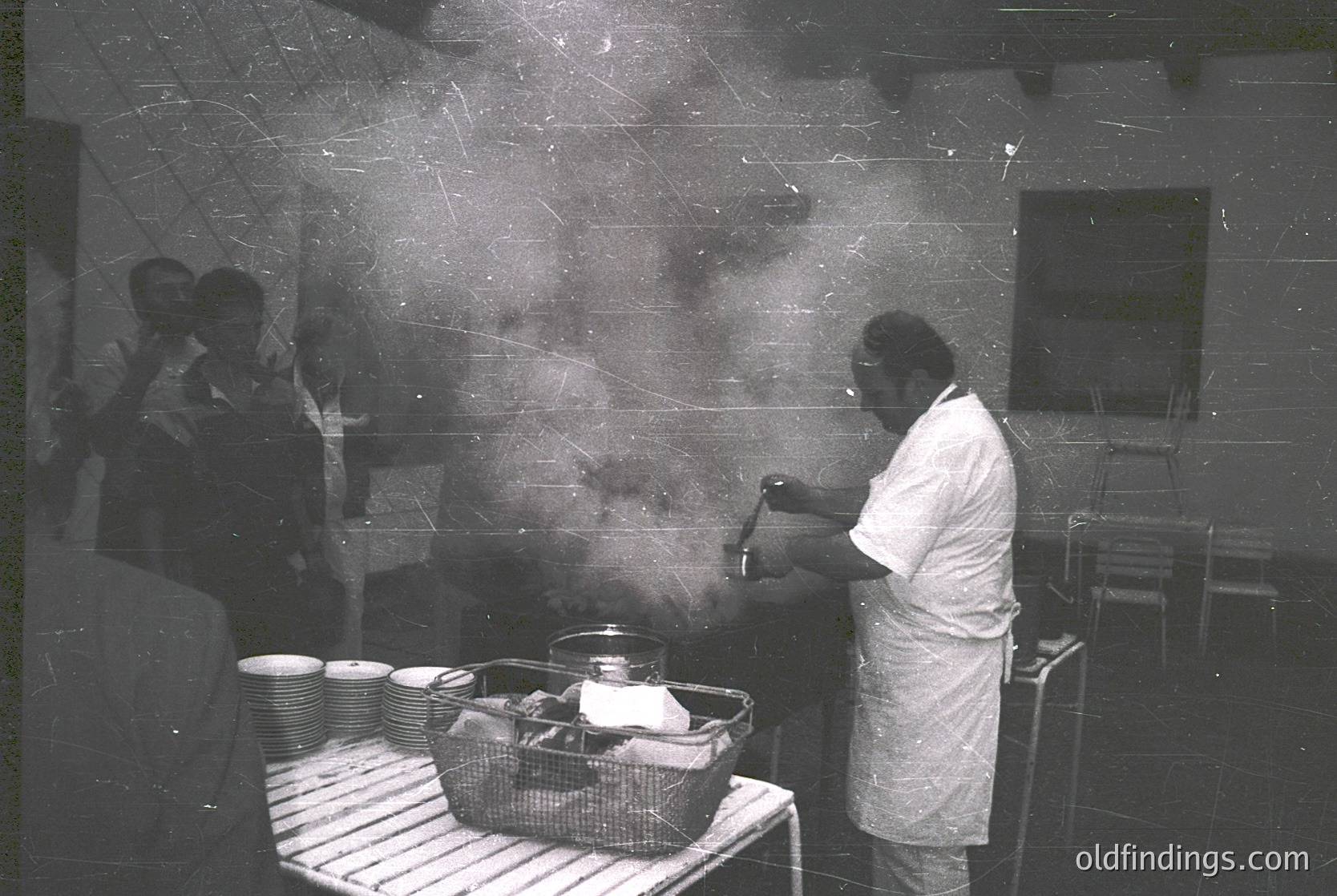 Black-and-white industrial kitchen scene from the 1960s–70s. Chef in white uniform ladles steaming liquid from a pot, surrounded by stacked white plates and a wicker basket. Industrial tiles and exposed pipes reflect mid-century work environment.