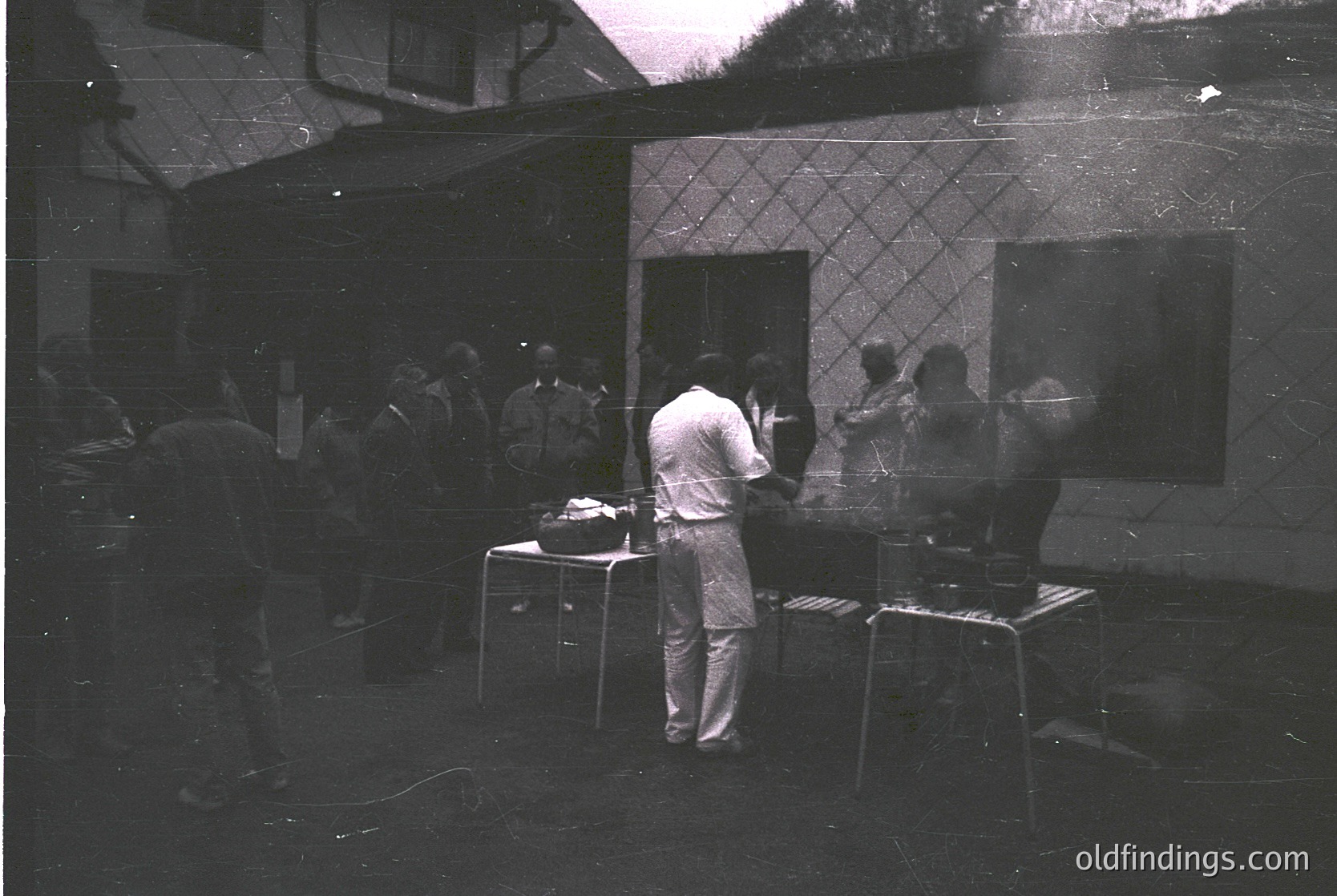 Vintage black-and-white photo of a small, dimly lit courtyard gathering, likely 1960s–1970s. A man in a white shirt and rolled-up pants stands at a table with a projector, casting light onto a patterned wall. Others sit on stools or lean against walls, observing. Minimalist urban setting with geometric tile accents.