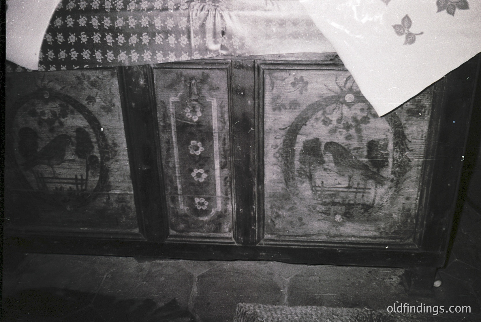 Vintage fresco-style wall panels featuring faded floral motifs and geometric designs, likely from a traditional Eastern European interior. Symmetrical arches and central floral medallions suggest 19th-century folk art influences. Stone base and partial patterned fabric above hint at preserved rural architecture.