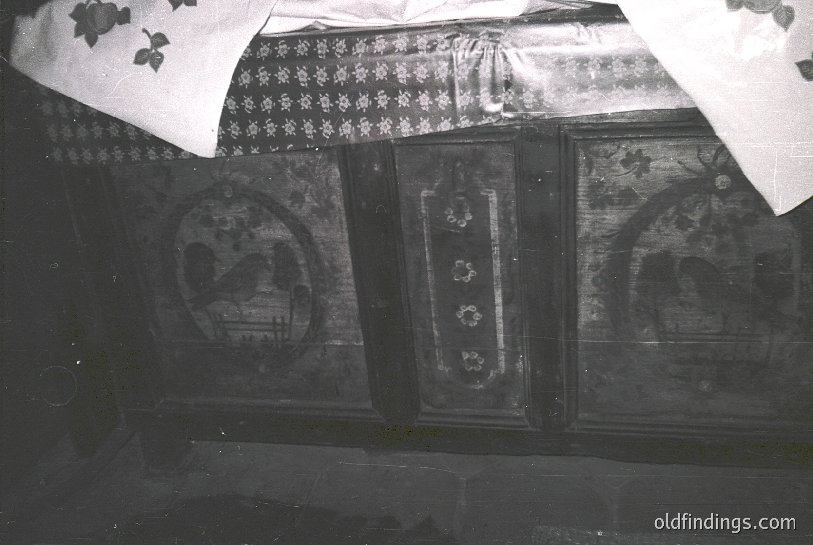 Vintage wooden cabinet with floral and geometric fresco-style panels, likely from a traditional Eastern European interior. Decorative embroidered fabric partially draped over the top, suggesting domestic use. Black-and-white photo indicates mid-20th century (1940s–1960s) craftsmanship.