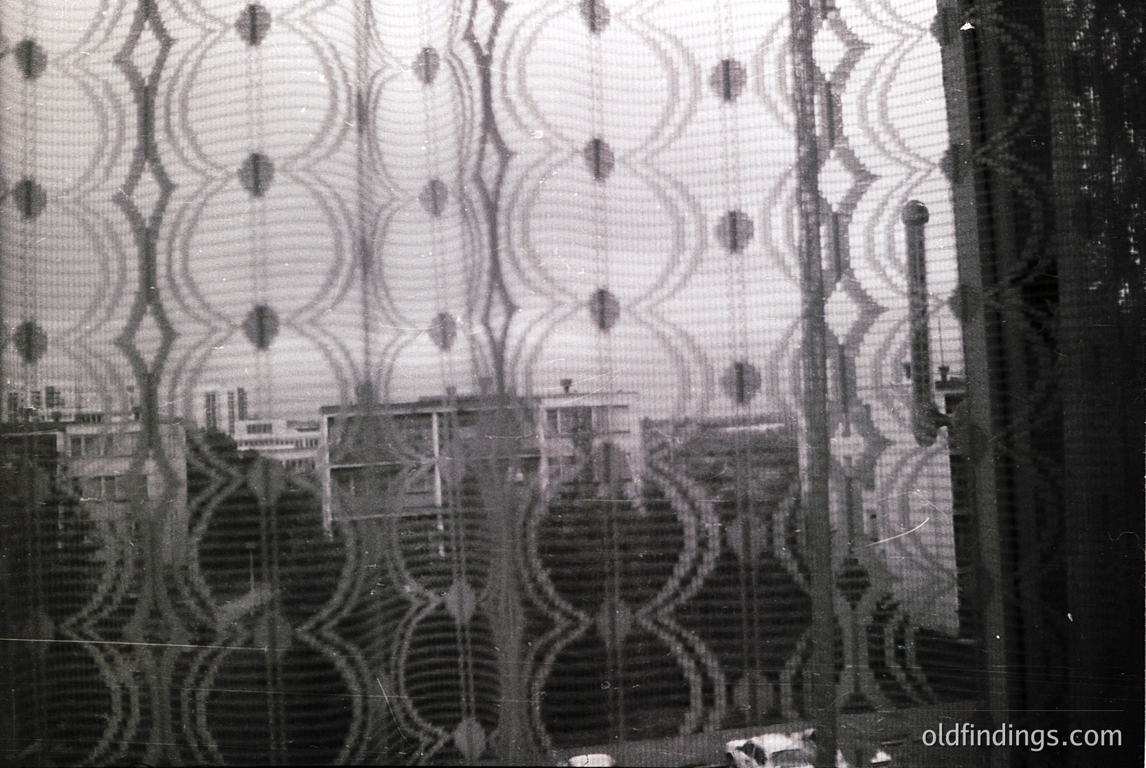 Distorted urban reflection through patterned glass, likely industrial or decorative. Symmetrical circular motifs create abstract cityscape distortions. Mid-20th century architectural elements visible behind glass.