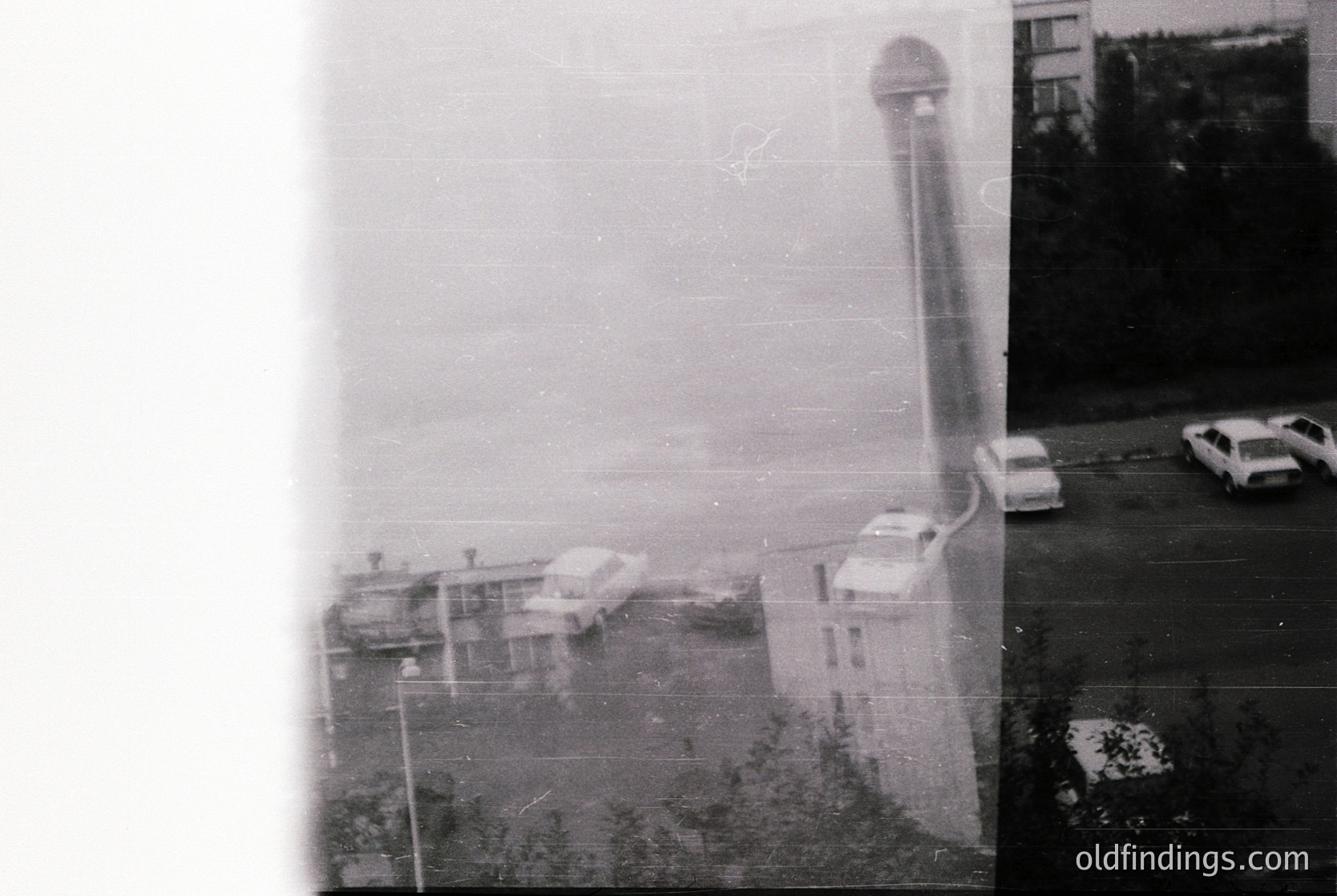 Vintage black-and-white shot of a coastal industrial site with a tall, cylindrical cooling tower and low-rise buildings. Fog obscures distant views, while parked cars and a fence frame the foreground. Likely Eastern Bloc-era infrastructure, 1960s–1980s.