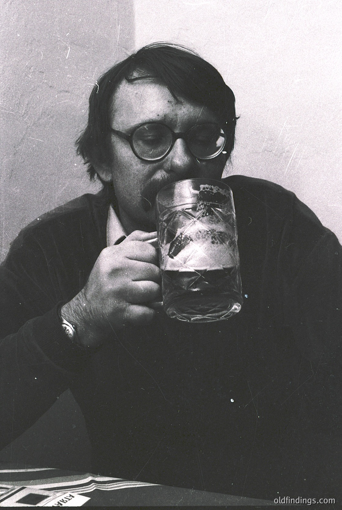 Mid-20th century man in round glasses sips from a frosted beer mug, likely in a café or pub setting. His attire suggests a formal-casual style, with a sweater and wristwatch. Black-and-white composition hints at 1950s–1970s vintage. Ideal for vintage lifestyle, historical research, or nostalgic design references.