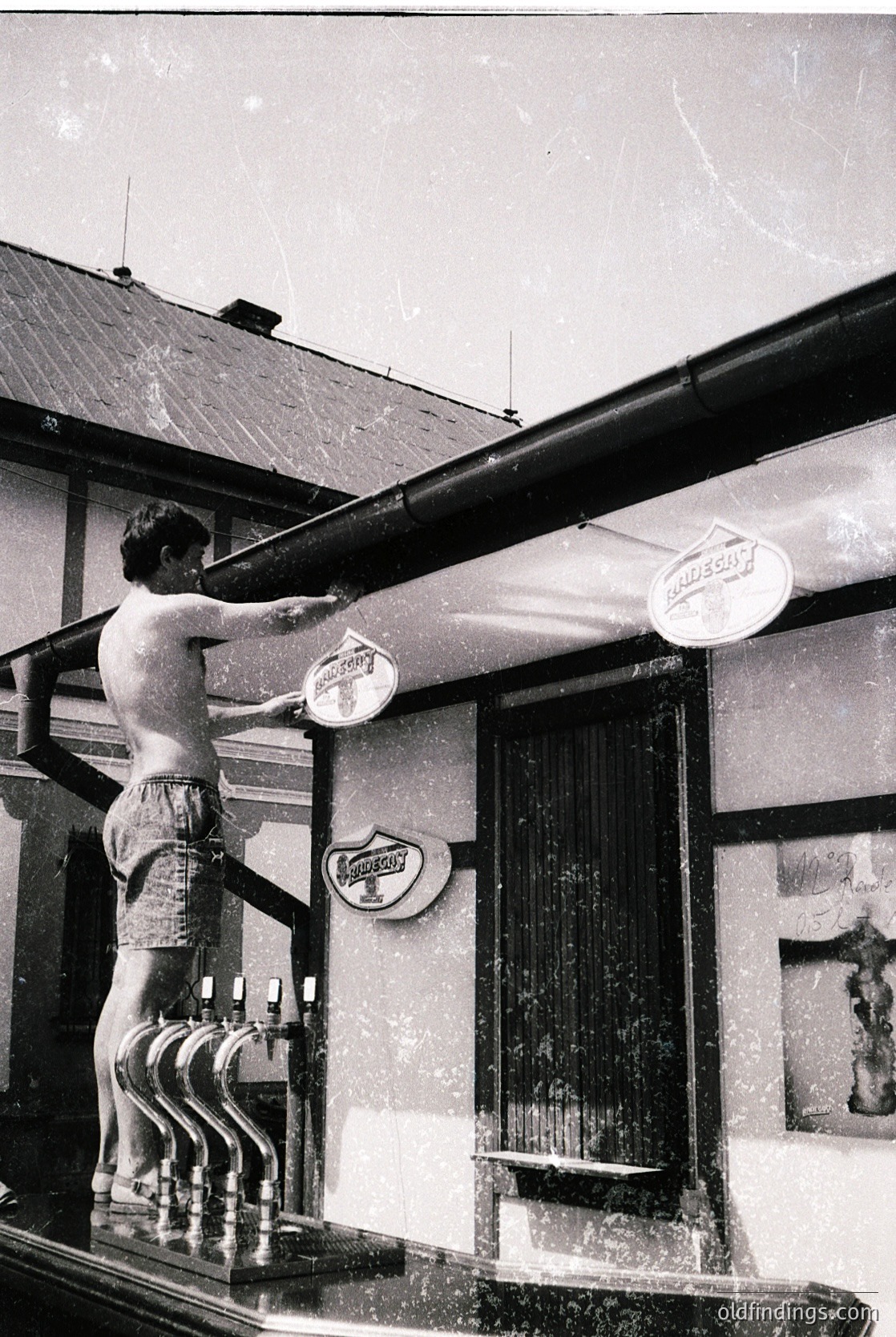 Mid-20th century black-and-white shot of a man in swim trunks washing a neon beer sign on a balcony railing. Wooden building with vertical blinds and vintage signage ( ).