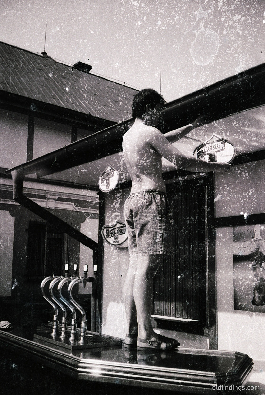 Vintage black-and-white photo of a man in swimwear cleaning a brass bar rail in a 1950s-60s American-style bar. Industrial-era fixtures, including a vintage soda fountain and hanging signs, suggest a retro diner or speakeasy setting.