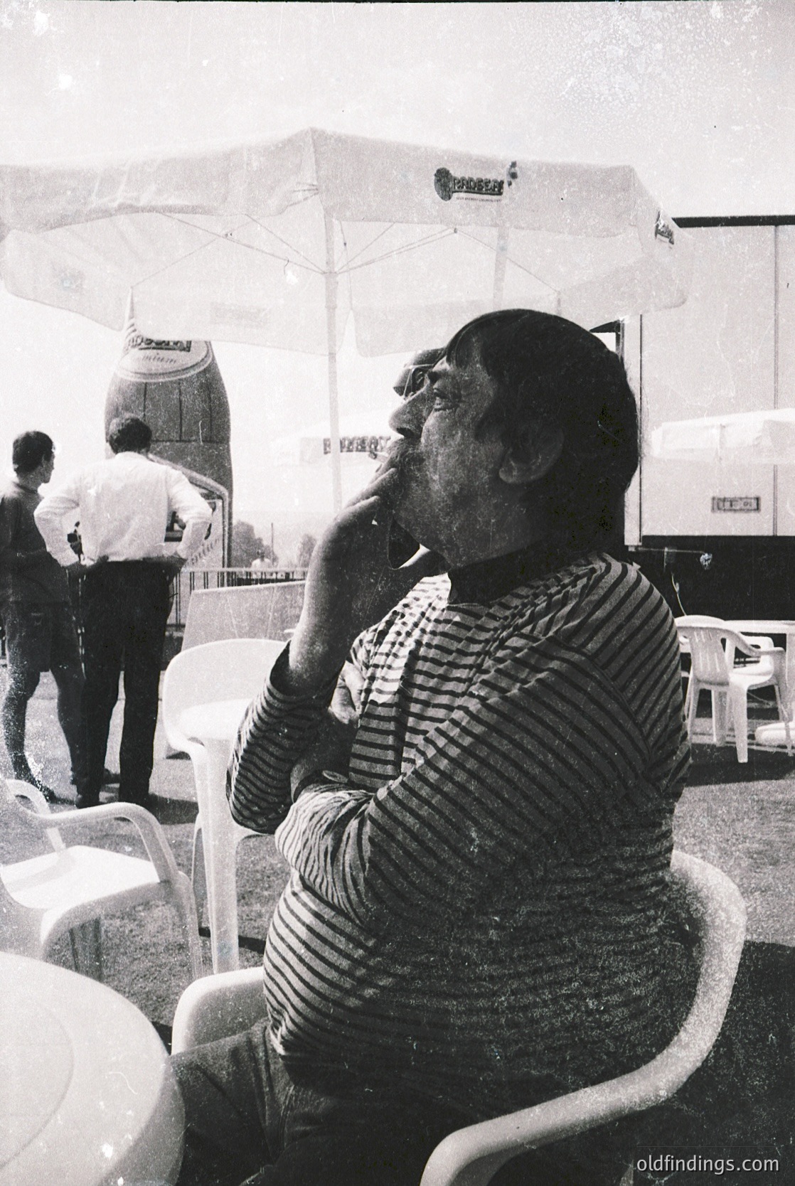 Mid-century outdoor café scene featuring a man in a striped sweater smoking, seated at a white plastic chair. Umbrellas and vintage signage suggest a retro European setting. Likely 1960s–1970s. é
