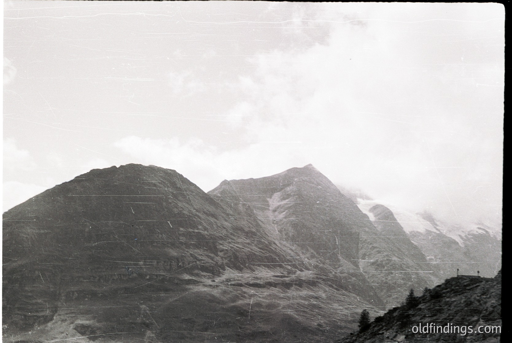Black-and-white alpine landscape featuring jagged peaks shrouded in mist, likely European Alps. Distinctive terraced slopes and sparse vegetation suggest early 20th-century mountaineering or agricultural use. Fog obscures mid-ground, emphasizing dramatic scale.