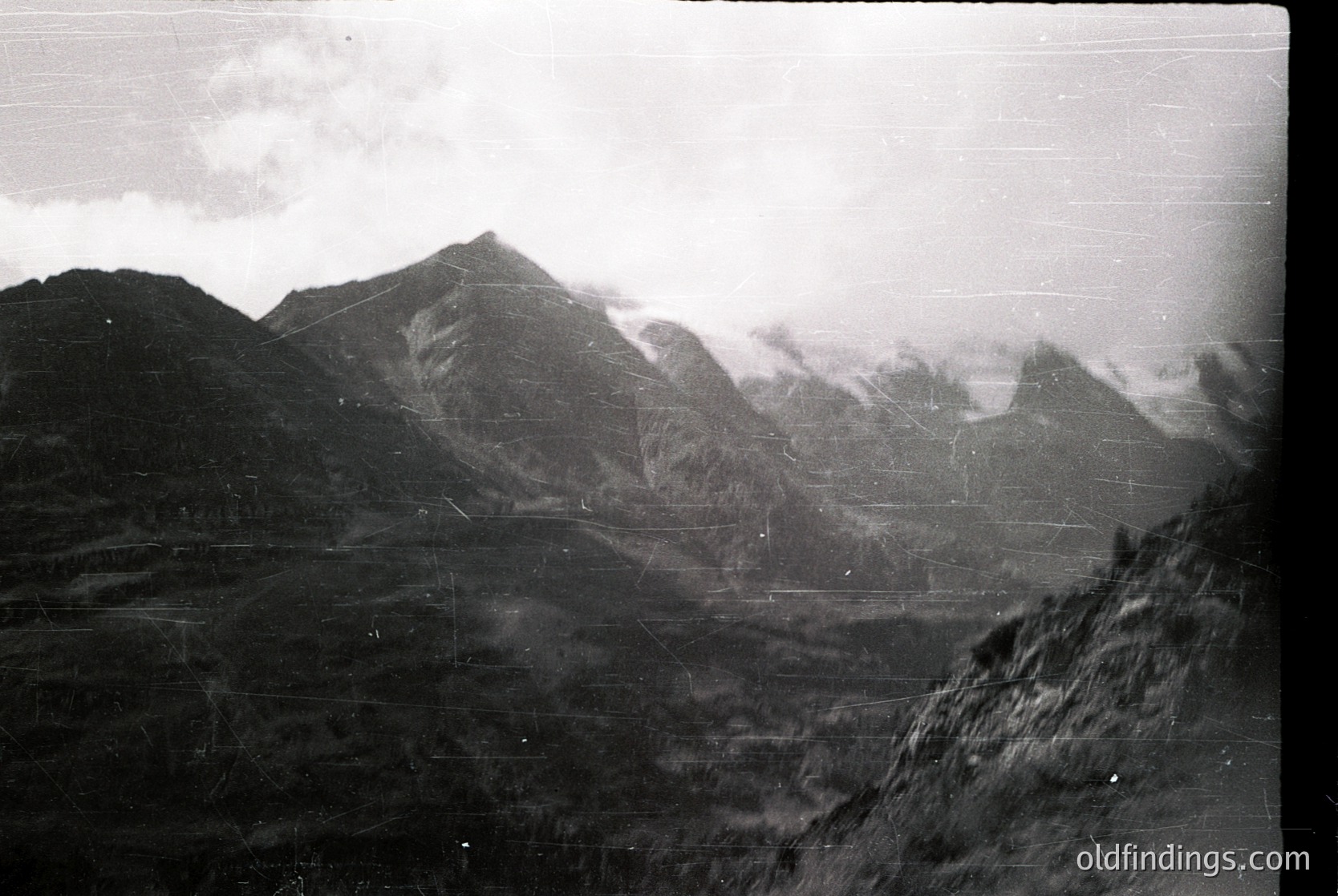 Vintage black-and-white alpine landscape with jagged peaks shrouded in mist, suggesting early 20th-century photography. Foreground shows rugged terrain with sparse vegetation. Film grain and slight damage enhance nostalgic vintage aesthetic.