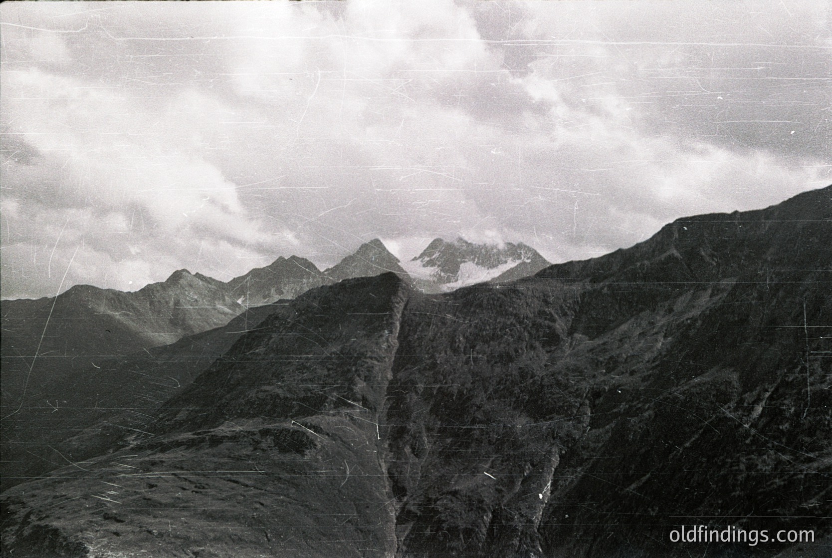 Vintage black-and-white aerial shot of rugged alpine peaks with jagged ridges, likely from the 20th century. Snow-capped summits contrast against dense forested slopes. Dramatic cloud cover enhances the scene’s raw, untouched beauty.