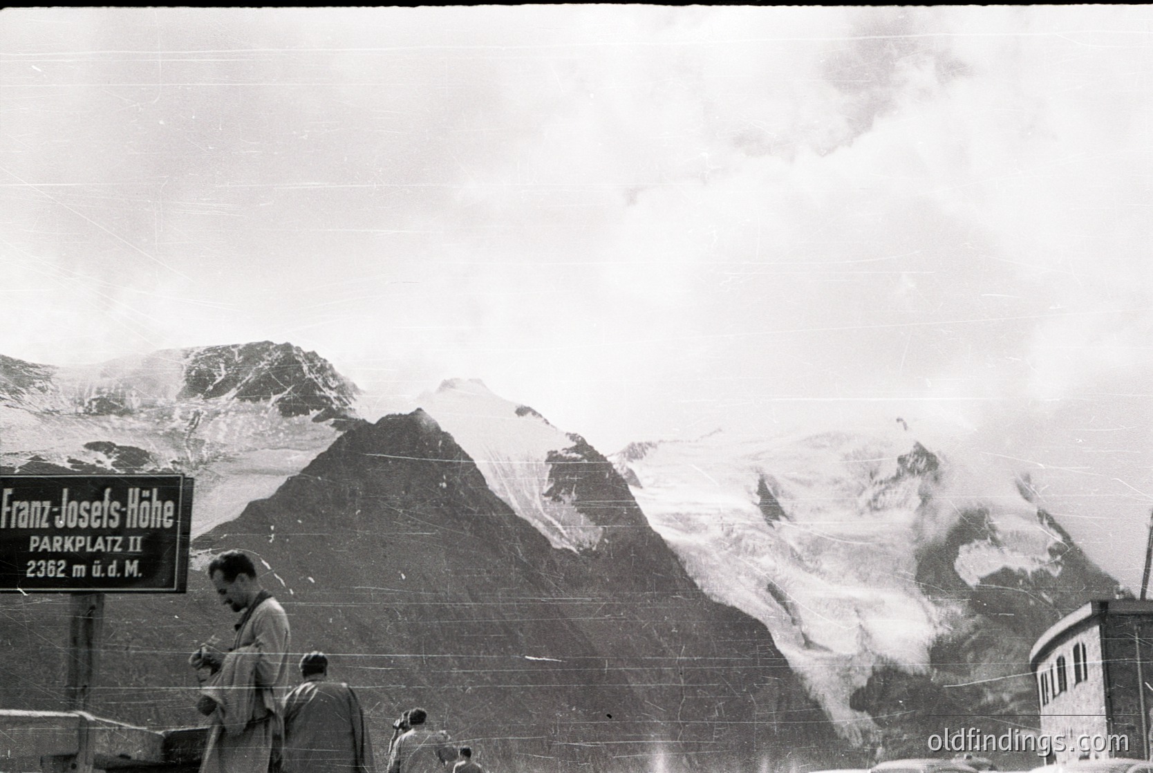 Black-and-white alpine scene at Franz-Josefs-Höhe (2392m) with sign indicating "Parkplatz II." Snow-capped peaks and misty valleys frame a small group of hikers in 1950s-era gear. Classic mid-century mountaineering aesthetic. öhe