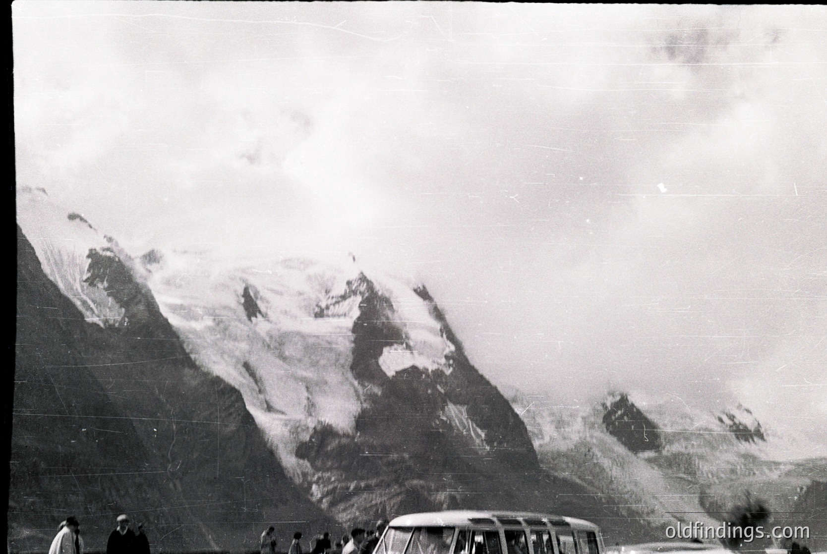 Snow-capped alpine peaks dominate this vintage black-and-white photo, likely from the mid-20th century. A vintage car and lone figure in foreground suggest a remote mountain pass or valley. Fog obscures mid-ground, enhancing dramatic scale.