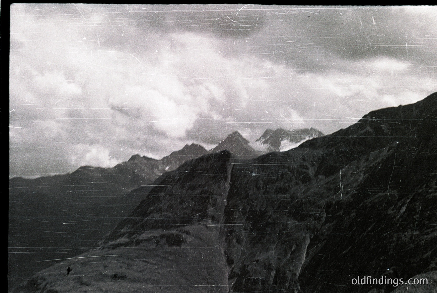 Vintage black-and-white alpine landscape with jagged peaks shrouded in mist, likely European Alps or similar range. Distinctive vertical scratches and film grain suggest mid-20th century photography. Dramatic lighting enhances rugged terrain and cloud formations.