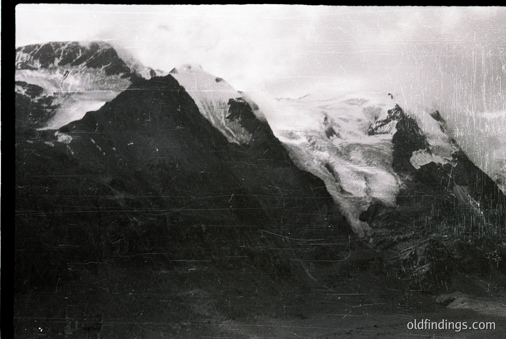 Vintage black-and-white alpine landscape with jagged peaks and snow-covered slopes, framed by a weathered, textured border. Dramatic cloud cover enhances the rugged terrain’s grandeur. Likely early-to-mid 20th century.