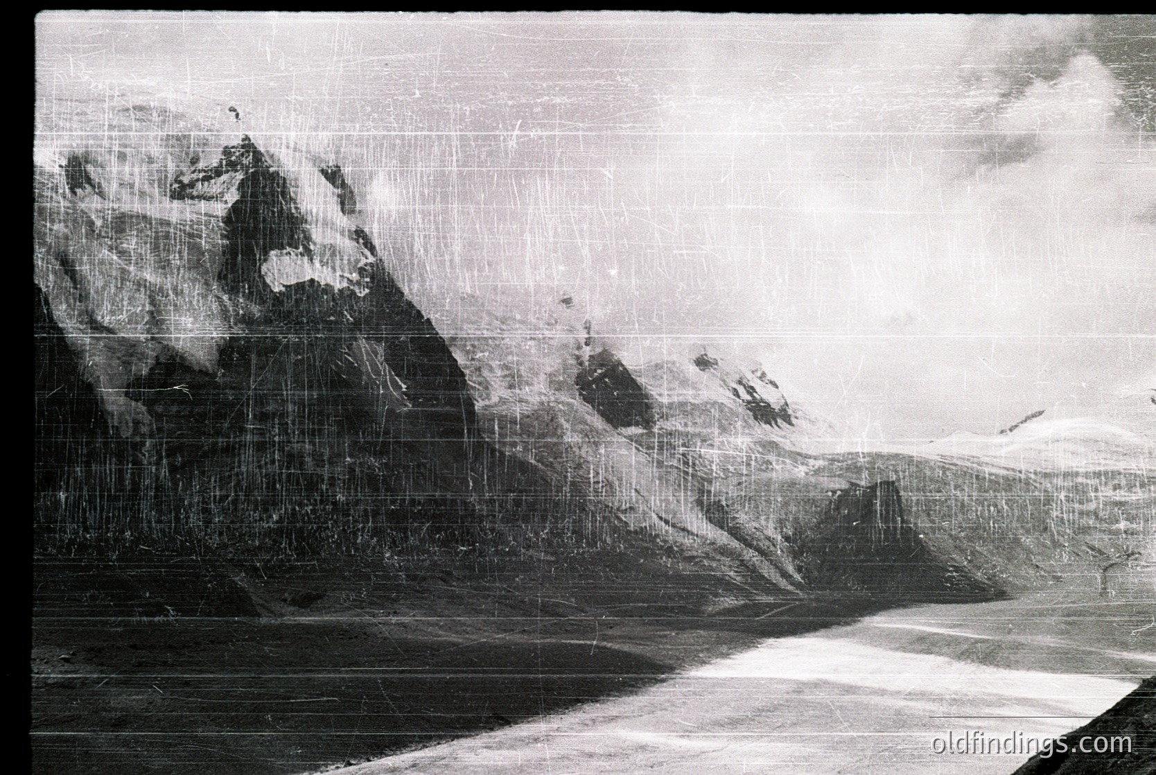 Vintage black-and-white aerial shot of rugged alpine terrain with jagged peaks and steep slopes, likely captured via early flight or balloon. Visible damage suggests aged film or print.
